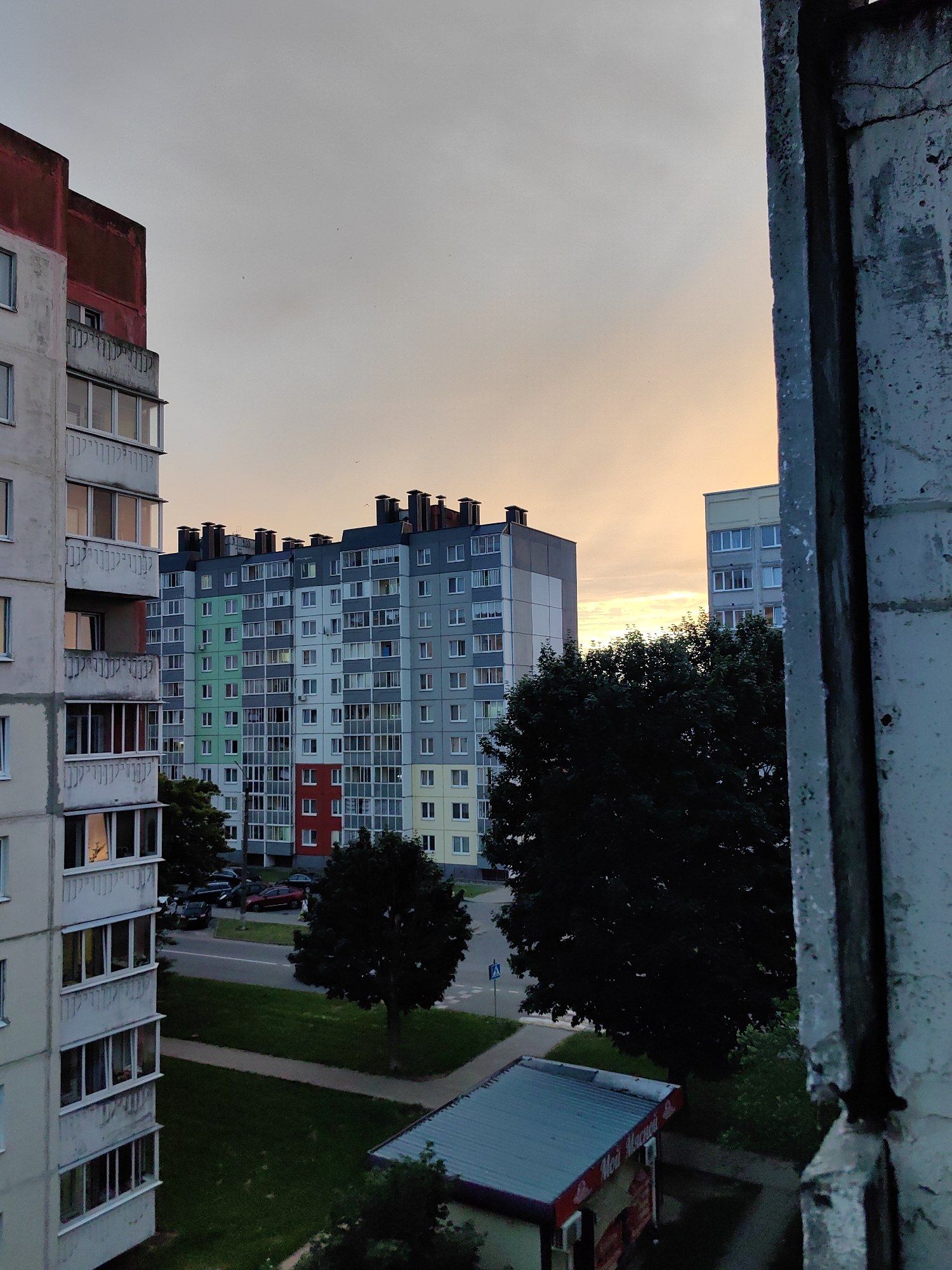 The image depicts a view from a window overlooking a residential area in Fanipal, Belarus. In the foreground, to the right, is a weathered concrete window frame. The middle ground features several apartment buildings. The building closest to the left is partially visible and appears to be under construction or renovation, with exposed concrete elements and a red-capped top section. To its right, a larger apartment building exhibits a colorful facade with sections of green, grey, red, and yellow. Another apartment building, mostly grey with yellow accents, is visible further in the distance, to the right of the colorful building. These buildings are surrounded by mature trees, green lawns, and a road with several parked cars. The sky above is a pale orange, indicating either sunrise or sunset, with a few faint birds visible. A small retail kiosk with a blue roof and signage in Cyrillic script is situated in the lower right portion of the scene. The overall impression is a view of urban life in the evening or early morning.
