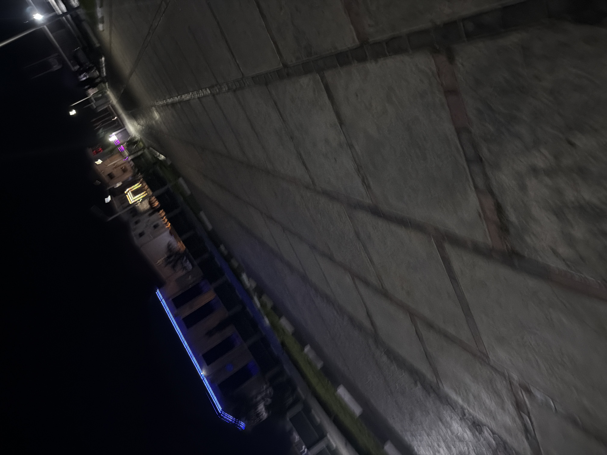 The image captures a street scene at night in Bukuru, Nigeria. The perspective is angled, looking down a paved walkway with a pattern of large, rectangular tiles. The walkway is bordered by a strip of grass and concrete bollards on the left side, leading towards a building illuminated by blue lights along its upper edge. The building appears to be a structure with multiple windows. Further down the street, other buildings are visible, some with decorative lighting, including a large rectangular outline of lights. Streetlights are also present, casting illumination on the scene. The overall impression is of a modern urban area at night, with artificial lighting playing a significant role in defining the visible elements.