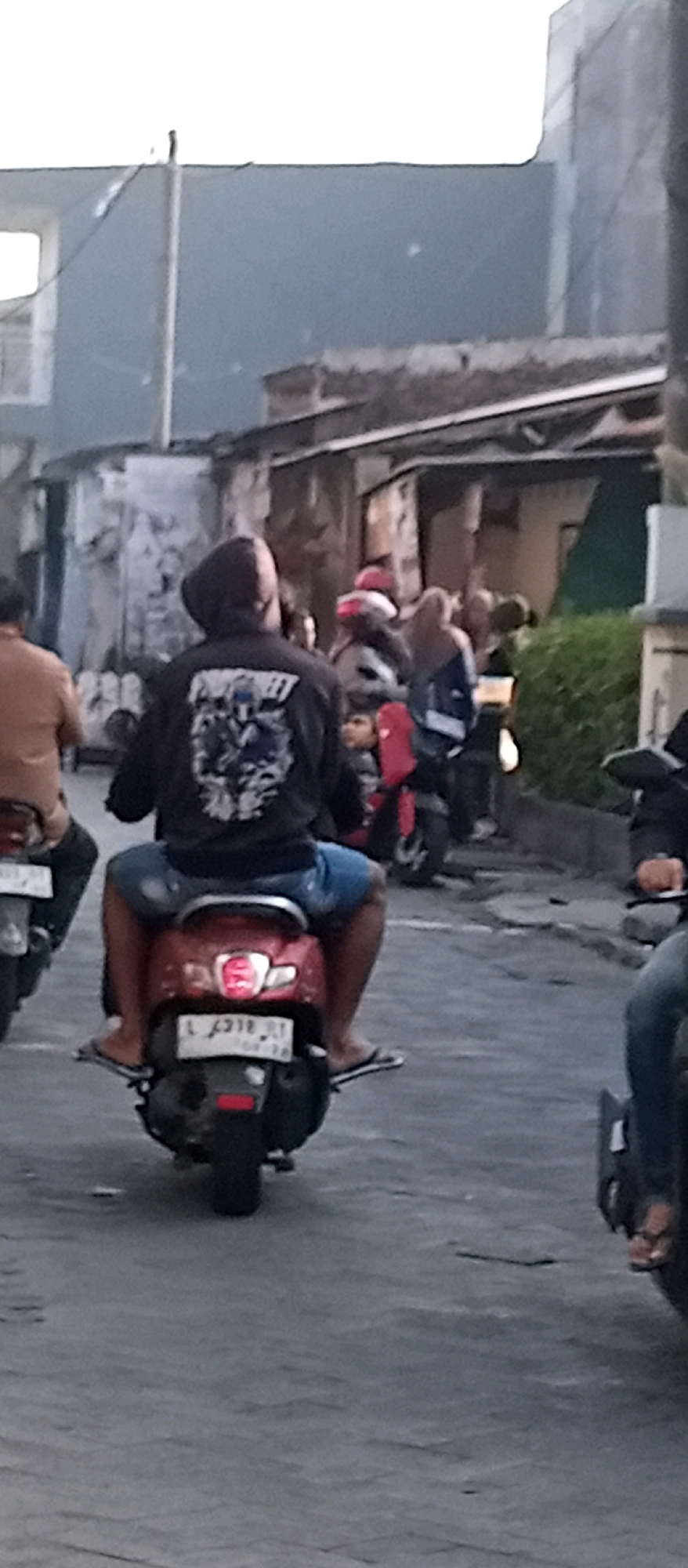 In Wadungasri, Indonesia, an individual wearing a black hooded sweatshirt with a graphic print on the back and blue denim shorts rides a red scooter. They are accompanied by a passenger also wearing blue denim shorts and possibly a white t-shirt, sitting behind them. The scooter has a license plate that reads "L 4918 JT" and has a red taillight. The background shows buildings with dilapidated walls and a street with other scooters and people. The lighting suggests it is either early morning or late afternoon.