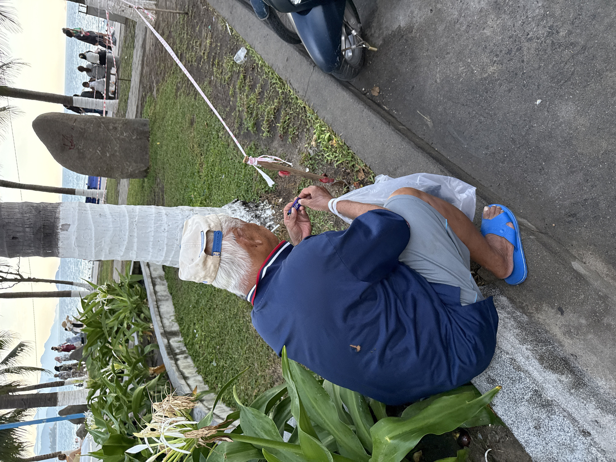An elderly Asian man wearing a blue polo shirt, light gray shorts, a white baseball cap, and blue flip-flops is squatting on a concrete curb. He is holding a small item, possibly a cigarette, in his right hand and a plastic bag in his left. He appears to be engaged in an activity related to the item he is holding. The background shows a coastal scene with palm trees, a body of water, and mountains in the distance. Several people are visible in the background, some sitting and others standing. A dark-colored motorcycle is parked to the right of the man. A pink and white caution tape is strung between poles in the mid-ground. The man's clothing has a small dark stain on the back of his shirt. The overall environment suggests an outdoor recreational area near the beach. The specific location is identified as Nha Trang Ward, Vietnam.