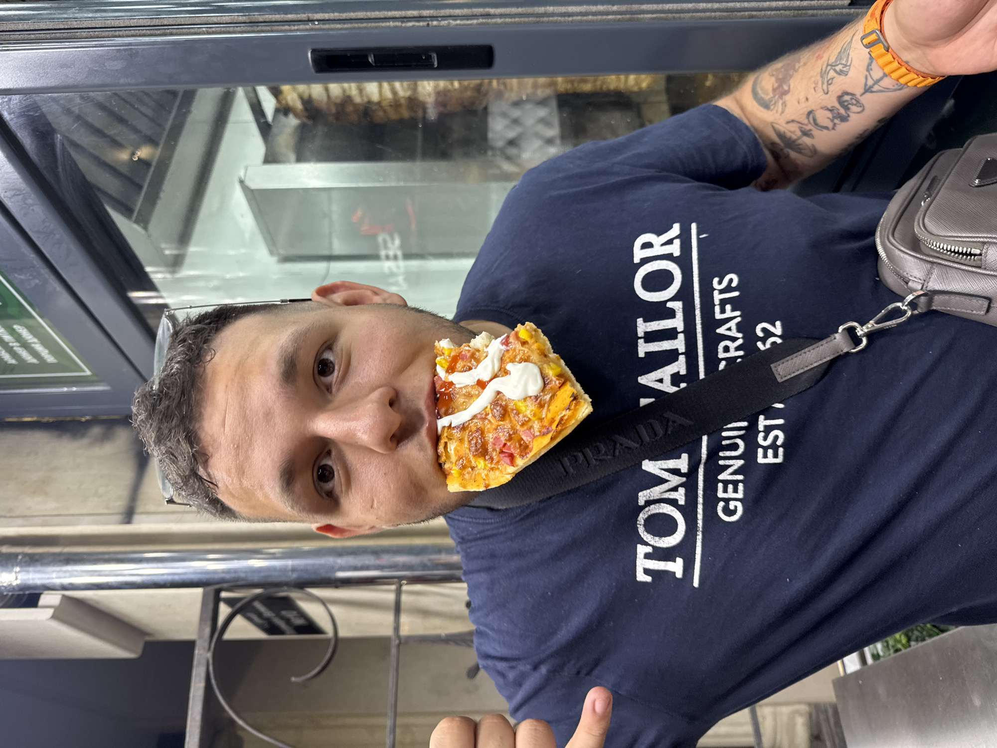A medium shot captures a man in a dark blue T-shirt with white text "TOM ' TAILOR GENUIN CRAFTS EST. 52" as he bites into a slice of pizza. He is wearing glasses on his head. A black strap runs across his chest, attached to a grey crossbody bag. His right arm, adorned with tattoos, sports an orange bracelet. He is standing in front of a glass-enclosed rotisserie with meat. The background includes a metallic pipe and a railing.