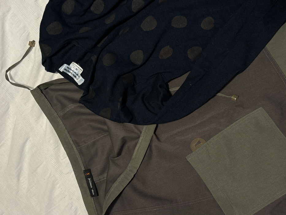 The image displays two articles of clothing arranged on a light-colored, textured fabric surface, likely a bed sheet or similar domestic textile. No specific location within the city of London, United Kingdom, is identifiable from the visual evidence.
The primary subject on the left is a garment, possibly a skirt or bottoms, in a dominant dark olive green or brown hue, featuring lighter olive green trim along the top edge and a rectangular patch pocket in the lower left quadrant. A drawstring, ending in a brass-colored metallic tip, is visible extending from the upper right. A black label with "cop. coping" and a stylized figure logo is sewn into the garment's interior near the waistband. Below the pocket, a circular, brass-colored metallic emblem is partially visible.
The secondary subject on the right is a dark blue or black long-sleeved top or cardigan, patterned with irregular, dark brown or black polka dots. A white rectangular label, bearing the text "COMME des GARÇONS COMME des GARÇONS" and indicating size "M", is affixed to the garment's neckline area.
The environment consists solely of the light, off-white, horizontally textured fabric on which the garments are laid, suggesting an indoor setting. No individuals, actions, or interactions are depicted.