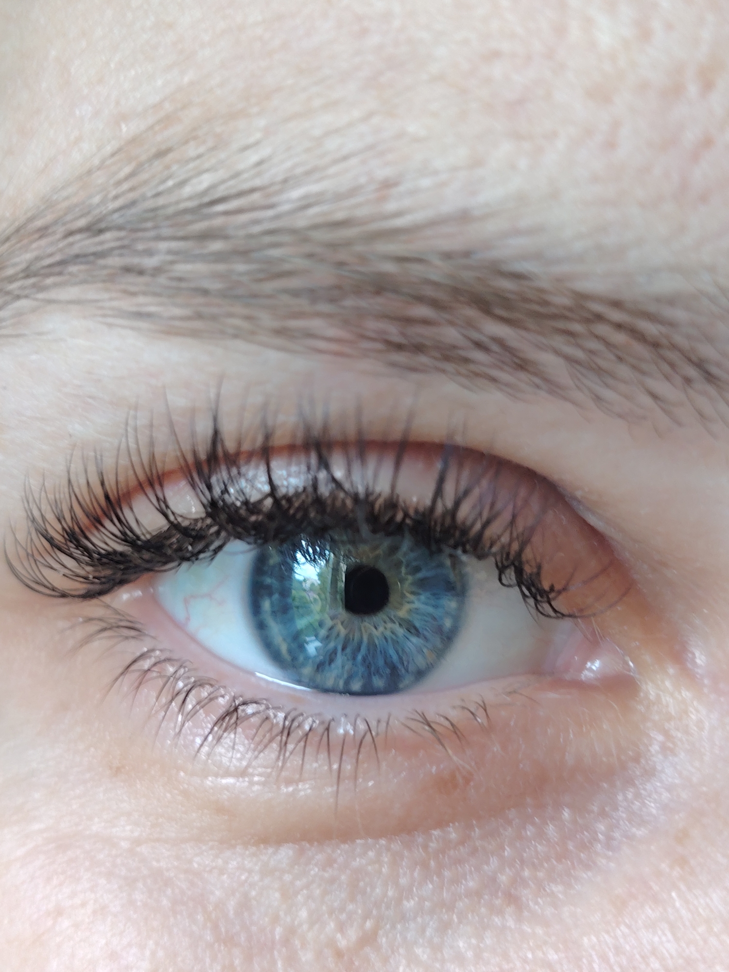 A detailed close-up of a human eye, located in the city of Chaikovsky, Russia. The prominent blue iris features a complex radial pattern of light and dark blue hues, originating from a perfectly round, dark pupil. Several bright reflections are visible within the pupil and iris, suggesting a window or outdoor light source, possibly reflecting green foliage. The upper eyelid displays numerous long, dark, and seemingly enhanced eyelashes, curling upwards. The lower eyelid has finer, shorter dark eyelashes. The white of the eye (sclera) shows faint red capillaries, particularly towards the inner corner. Above the eye, a portion of a dark, natural eyebrow with individual hair strands is visible. The surrounding skin is fair-toned, exhibiting fine texture and pores, with slight creasing near the outer corner of the eye.