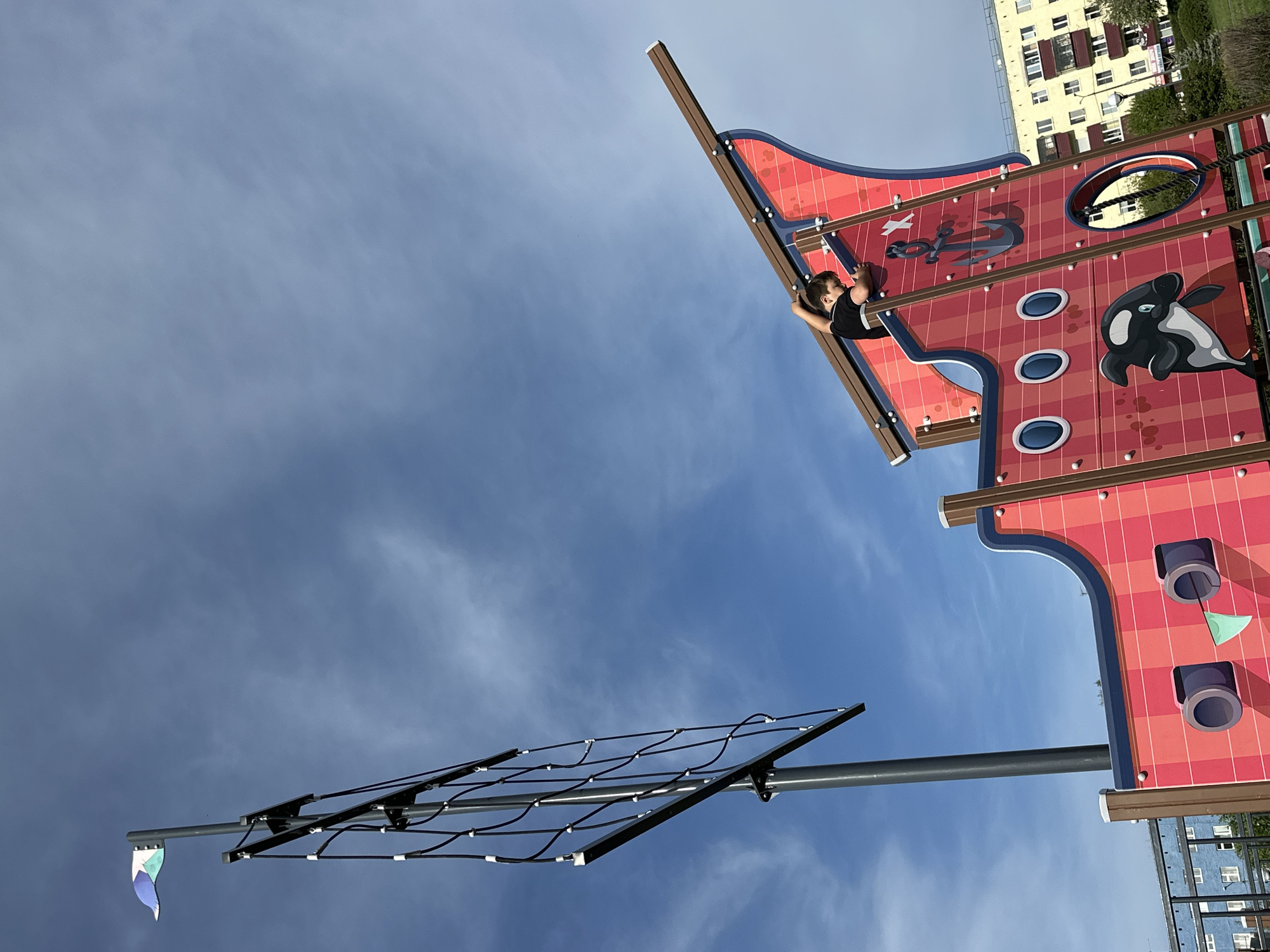 The photograph depicts a playground structure in Unknown, Unknown. A child in a black shirt is on the structure, which is designed to resemble a ship, adorned with nautical imagery, including an orca and an anchor. A climbing net stands on a pole. The sky is blue with scattered clouds, and a multistory building is visible in the background.