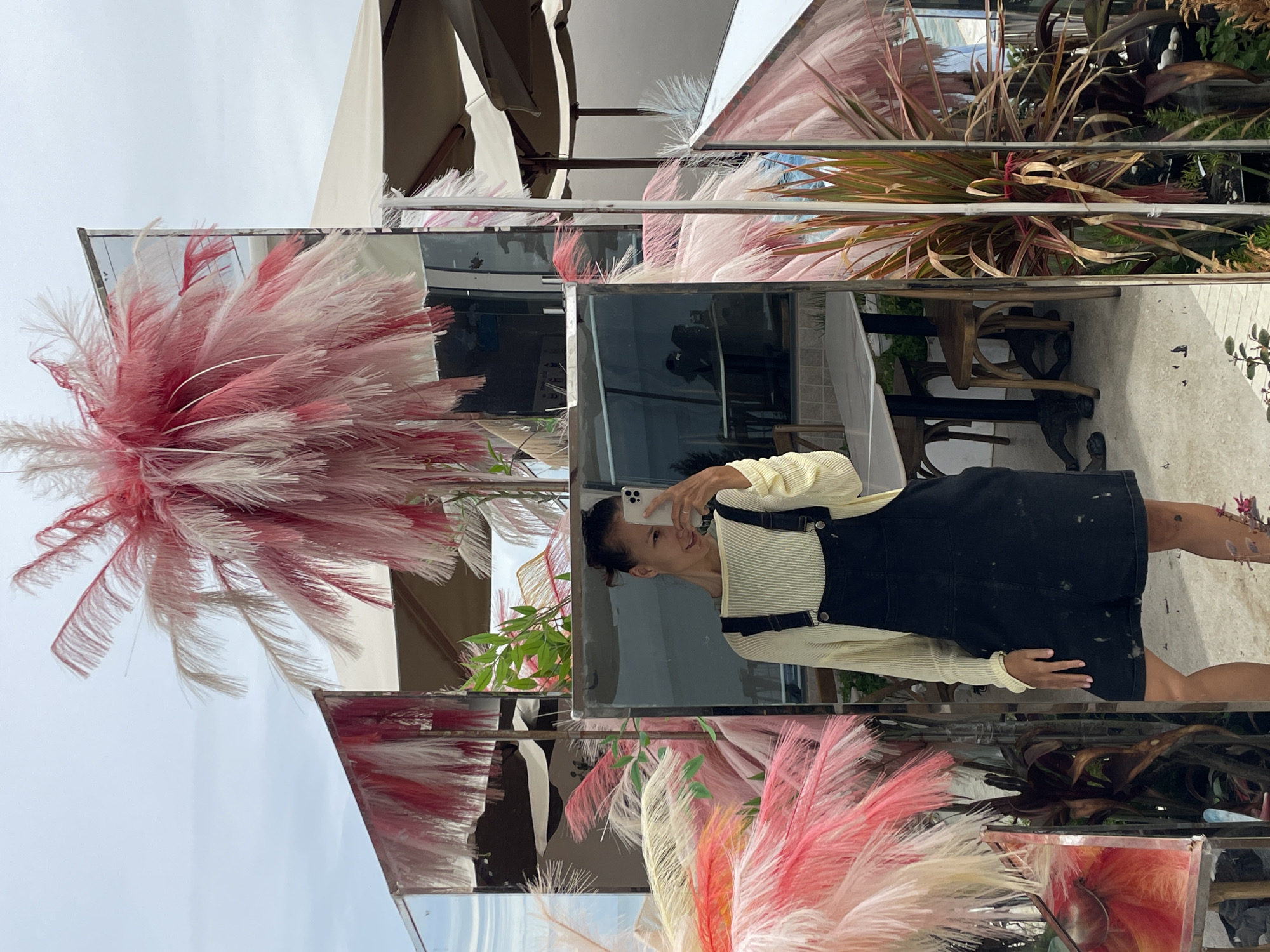 A woman in a cream-colored sweater and black overalls is taking a selfie in a mirrored structure. The mirrors reflect pink and white feathered decorations, greenery, and outdoor seating. The location is Phu Thuy Ward, Vietnam.