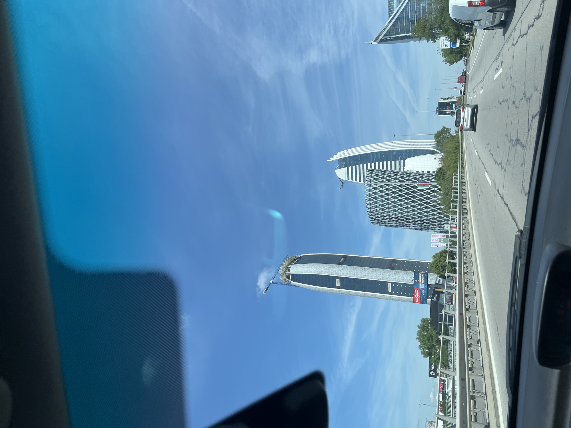 The image shows a view from inside a vehicle on a highway in Sofia, Bulgaria, looking towards a cluster of modern skyscrapers. The skyscrapers are characterized by their distinct architectural designs, with one being a tall, slender tower with a curved facade and a crane at its apex, indicating ongoing construction. Another prominent building features a diamond-shaped lattice facade. In the foreground, the highway is visible with several cars and a van. The sky is blue with scattered clouds, and there are trees and other buildings along the roadside. Advertisements and signs are visible on billboards and buildings, including one for Renault. The overall scene depicts an urban environment with contemporary architecture.
