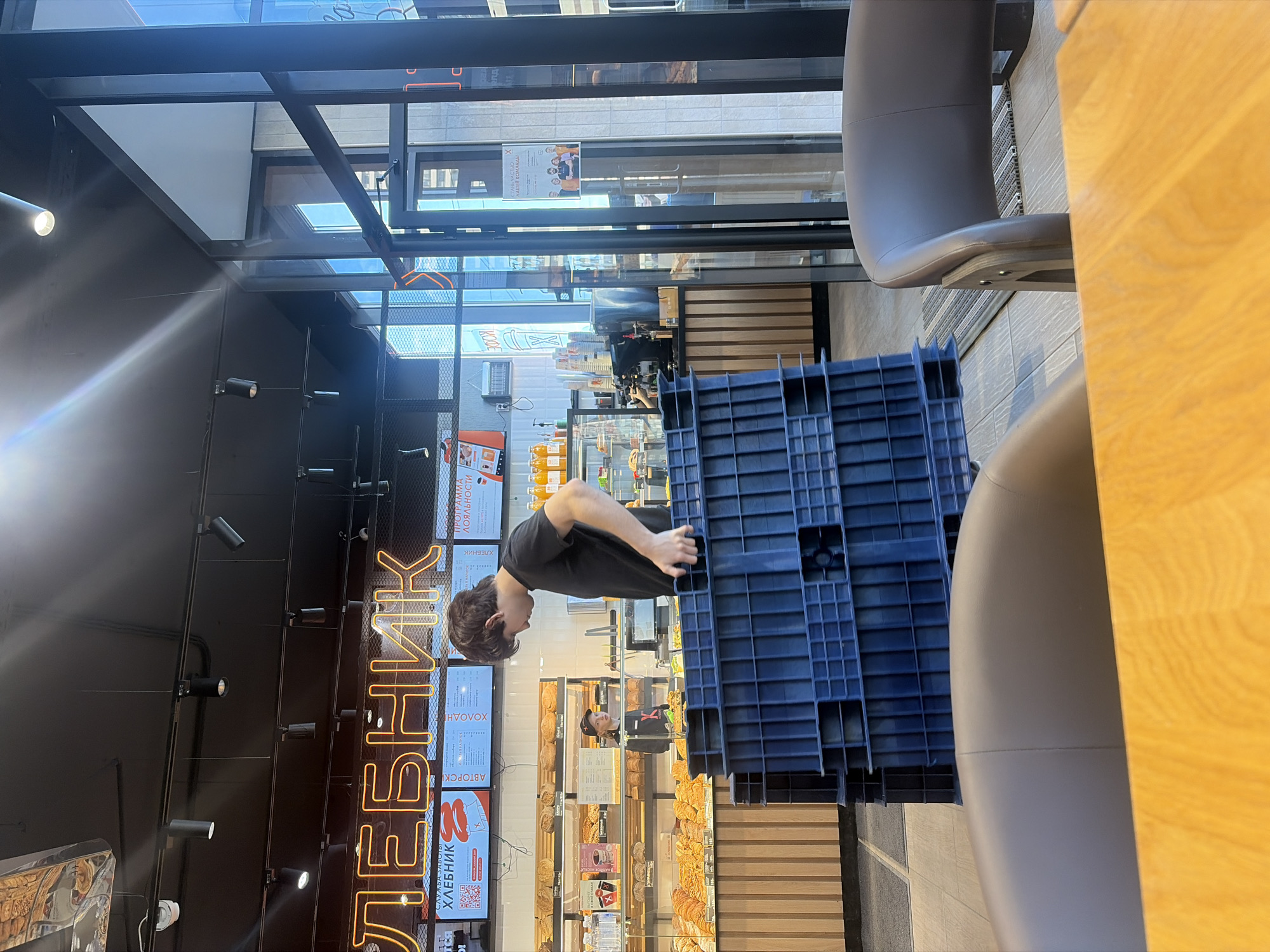 The image displays the interior of a ХЛЕБНИК (Khlebnik) bakery and cafe, a known chain operating in Saint Petersburg, Russia. In the mid-ground, a young male, likely in his late teens or early twenties, with short dark hair and wearing a black t-shirt, is depicted carrying a stack of four dark blue, rectangular plastic crates. He is oriented left-to-right, slightly bent forward, with his hands gripping the crates.
Behind him, to the left, a female employee in a dark uniform with a light baseball cap is visible behind a service counter, looking towards the front of the establishment. The counter features multiple tiers displaying a variety of baked goods, including loaves of bread, croissants, and pastries. Above the counter, illuminated digital screens display menu items and promotions, beneath a prominent orange neon sign spelling out "ХЛЕБНИК" in Cyrillic. Bottles of juice and coffee-making equipment are visible on a section of the counter to the far right.
The ceiling is black, fitted with black track lighting and multiple spotlights. Large, floor-to-ceiling glass windows and a glass-paneled entry door with black frames are situated on the right side of the image, allowing natural daylight to illuminate the space. A white sign with Russian text and an image of a group of people is affixed to the glass door. The floor is tiled. In the foreground, the upper backs of two brown leather-upholstered chairs and a portion of a light wooden table are visible.