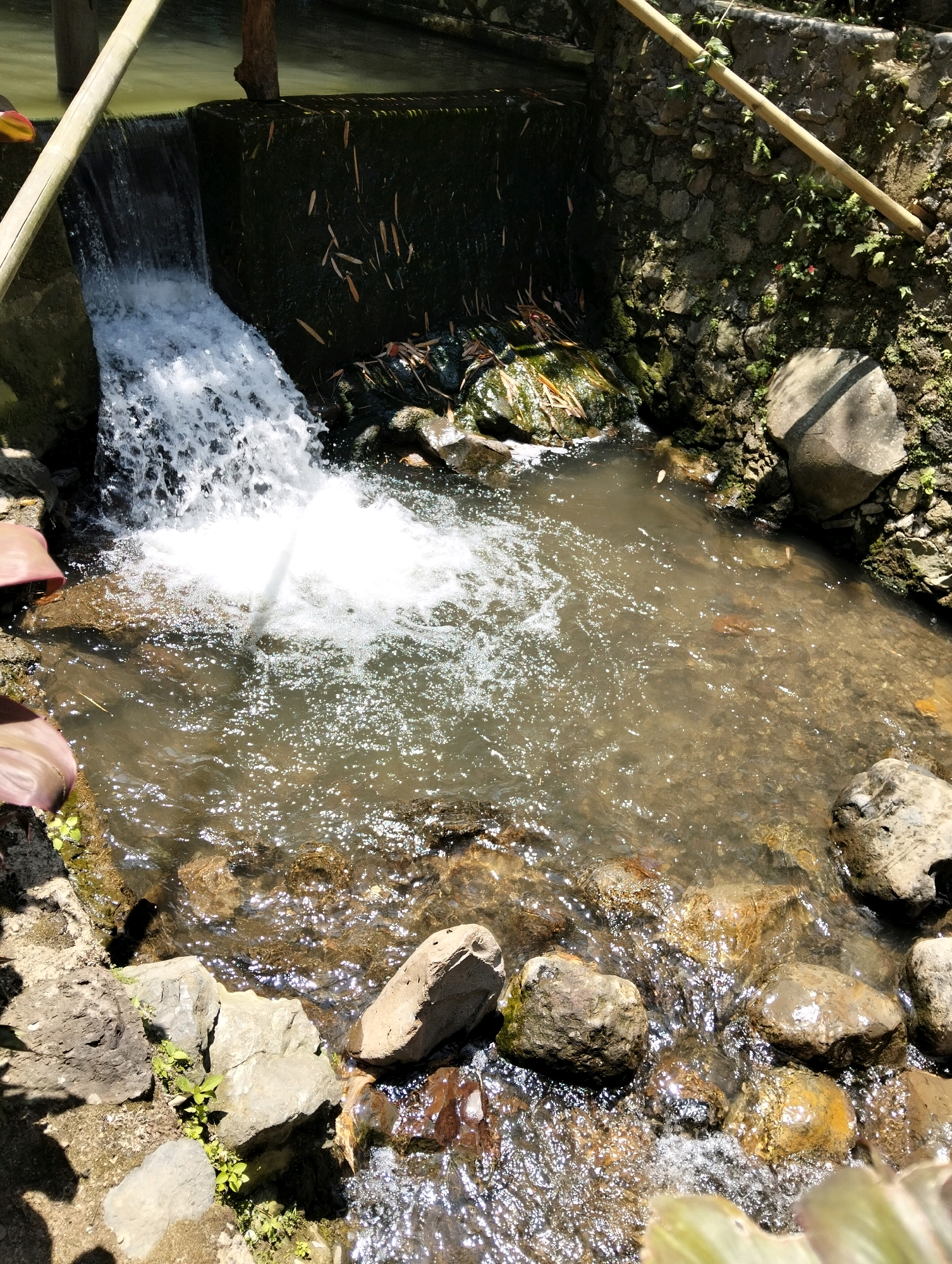 A small waterfall cascades into a shallow, rocky stream in a rural or natural setting, possibly in Subang, Indonesia. The water is clear with sunlight sparkling on its surface. The stream bed is comprised of various sized rocks, some smooth and rounded, others angular and rough. Green vegetation, including moss and small plants, grows on and around the rocks and along the banks. A dark, rough-textured wall, possibly made of stone or concrete, forms the edge of the waterfall, with some dried leaves and debris visible on it. A bamboo pole rests diagonally across the top of the scene, partially obscuring the view of the water source. The surrounding environment appears to be natural or semi-natural, with shadows suggesting it is daytime.