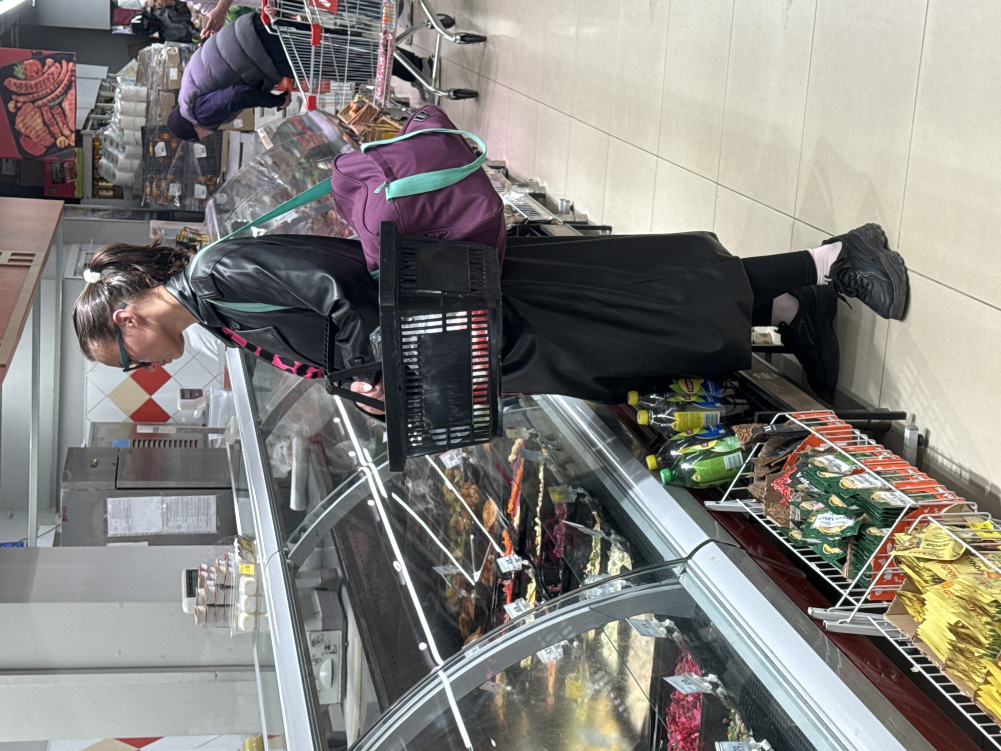 This image captures a scene inside a supermarket or grocery store in Yekaterinburg, Russia, depicting two individuals engaged in shopping.
In the foreground, a woman with dark hair, pulled back with a white scrunchie, is seen wearing black glasses, a black leather-like jacket, black leggings, and black chunky sneakers with light pink socks. She holds a small black plastic shopping basket in her left hand and carries a purple backpack with a prominent mint green strap. She is looking down into a refrigerated display case, likely inspecting or selecting an item.
Further down the aisle, another person is partially visible, pushing a metal shopping cart. This individual is wearing a dark purple or grey quilted vest.
The store environment features bright interior lighting and a light-colored tiled floor. A row of refrigerated display cases runs along the aisle, showcasing various food products. The case directly in front of the foreground woman appears to contain packaged meats or sausages, indicated by small white price tags such as "37" and "69." Above these cases, shelves are stocked with assorted packaged goods. Visible items include bottles of Lipton iced tea and racks filled with sachets of condiments. Text on these sachets is in Cyrillic, with "МАЙОНЕЗНЫЙ" (Mayonnaise) and "РУССКАЯ ГОРЧИЦА" (Russian Mustard) being discernible, reinforcing the Russian location context. In the background, a large display of raw meat or sausage products is visible, and further back, a section of wall with red and white diamond-patterned tiles can be seen near a grey counter with a scale. The overall atmosphere is that of a functional, busy retail environment during what appears to be daytime.