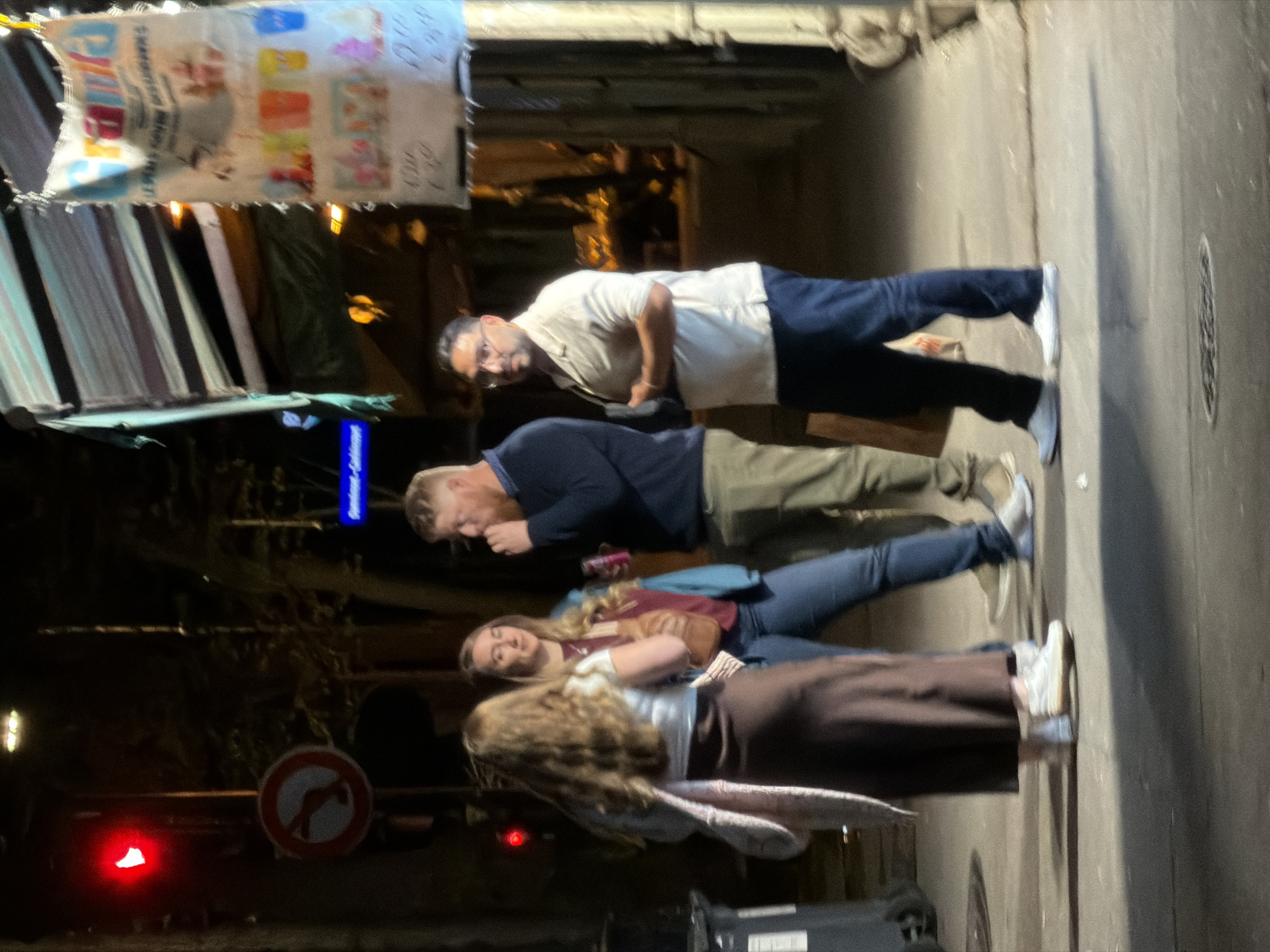 Four people are standing on a sidewalk next to a street in Paris, France. Overhead is a striped awning and a sign with faded lettering and images. The person nearest to the camera has long blonde hair and is wearing a white shirt and long brown skirt. The person behind them is blonde and wearing jeans. Behind them is a man in olive-colored pants and a blue shirt. Behind him is a man in blue pants and a white shirt. A "no U-turn" traffic sign is visible.