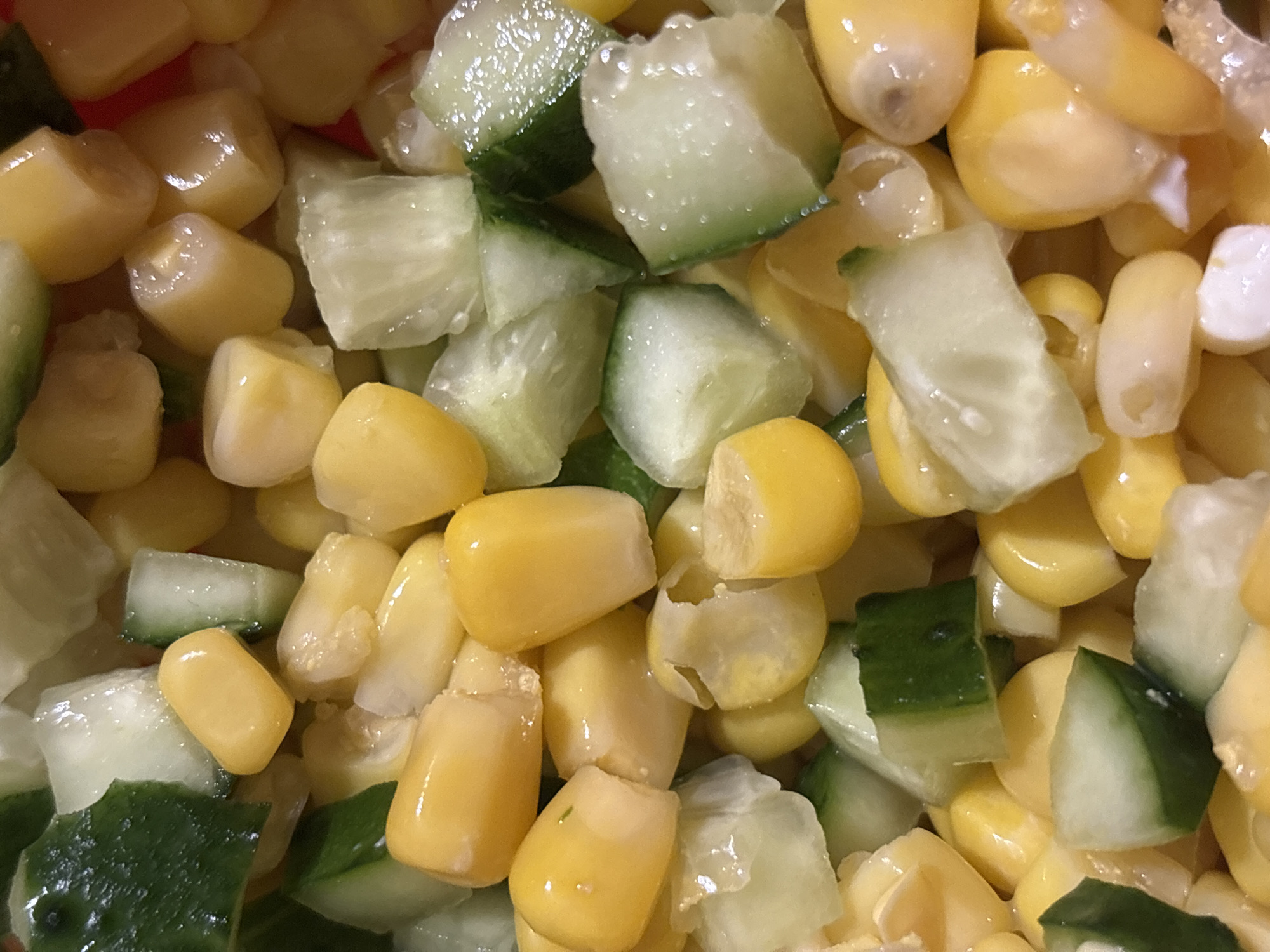 A close-up, overhead shot shows a salad composed of corn kernels and diced cucumber. The corn is bright yellow, with some kernels appearing plump and others slightly shriveled. The cucumber pieces are cut into irregular cubes, with visible green skin on some. The salad appears moist, suggesting it may have been dressed. The lighting is bright, casting highlights on the wet surfaces of the ingredients. The background is not visible, as the image is tightly cropped on the salad. The location in the city of Kremenchuk, Ukraine, is not determinable from the visual information provided.