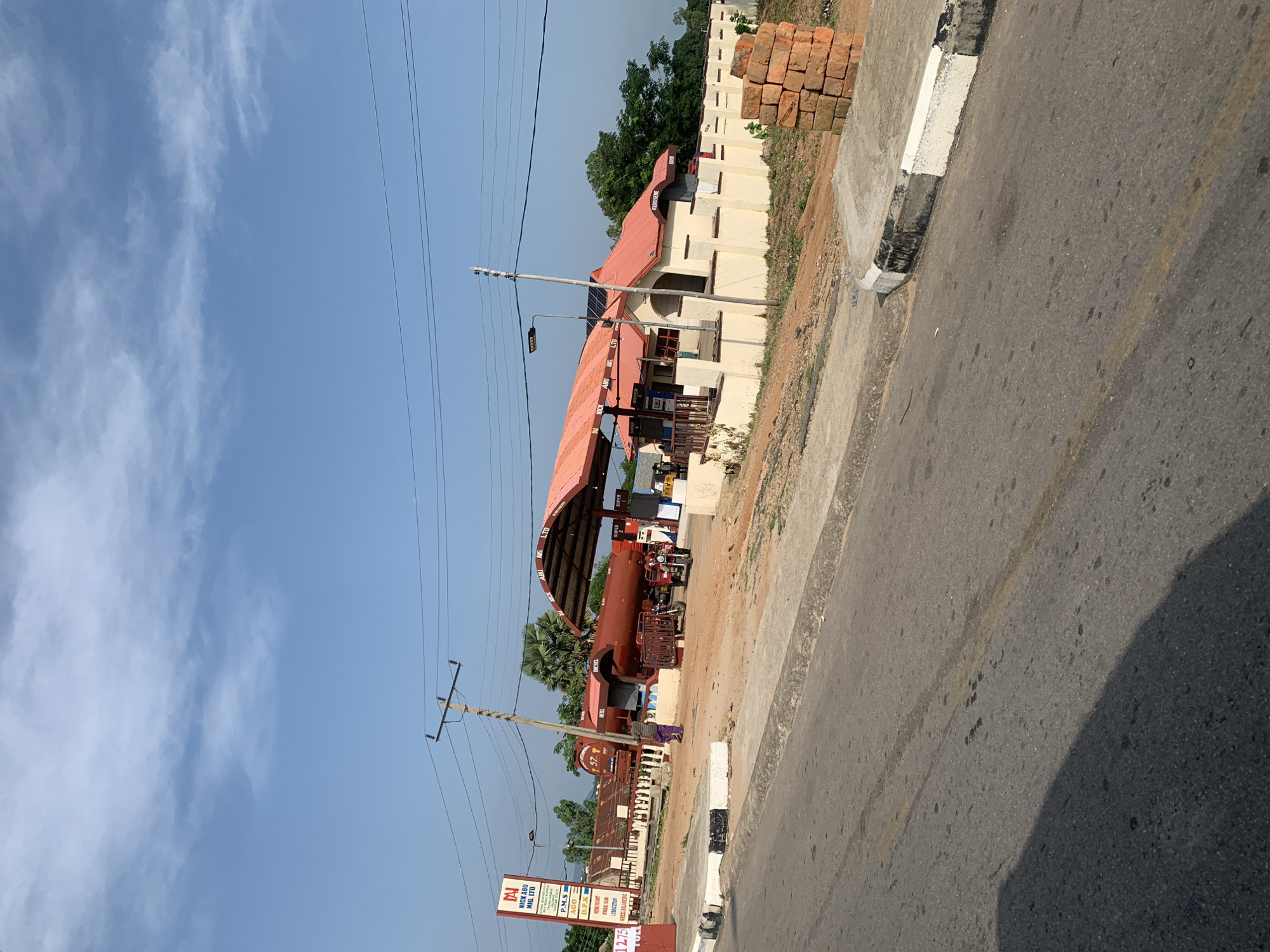 The image depicts a gas station in Jalingo, Nigeria, on a bright, clear day with a blue sky and scattered white clouds. The gas station, identified by a sign as "NICK ABU NIG. LTD," features a prominent red roof covering the pumps. A large, red, cylindrical fuel tanker truck is parked behind the station, partially obscured by a fence. Several vehicles, including what appears to be a motorcycle-drawn cart and a small truck, are present. People are seen near the station, with one individual walking by the fuel tanker. The foreground shows a paved road with a yellow line down the center and a concrete curb. A stack of bricks is visible on the right side of the image. The overall environment suggests a somewhat rural or semi-urban setting.