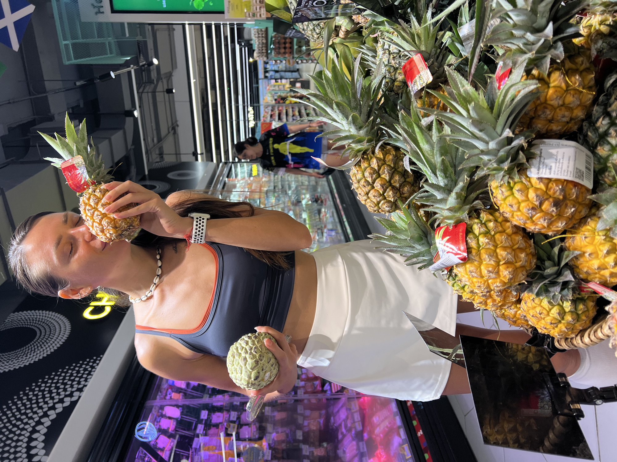 In Baan Chaweng, Thailand, a woman is captured in a supermarket aisle, near a display of pineapples. She's wearing a dark sports bra with orange trim and a white skirt. The woman is holding a small pineapple to her face, seemingly smelling it, and in her other hand, she holds a cherimoya fruit. Behind her, a blurred figure walks down the aisle. The supermarket environment is visible with shelves of products and overhead lighting. A price tag is visible on one of the pineapples.