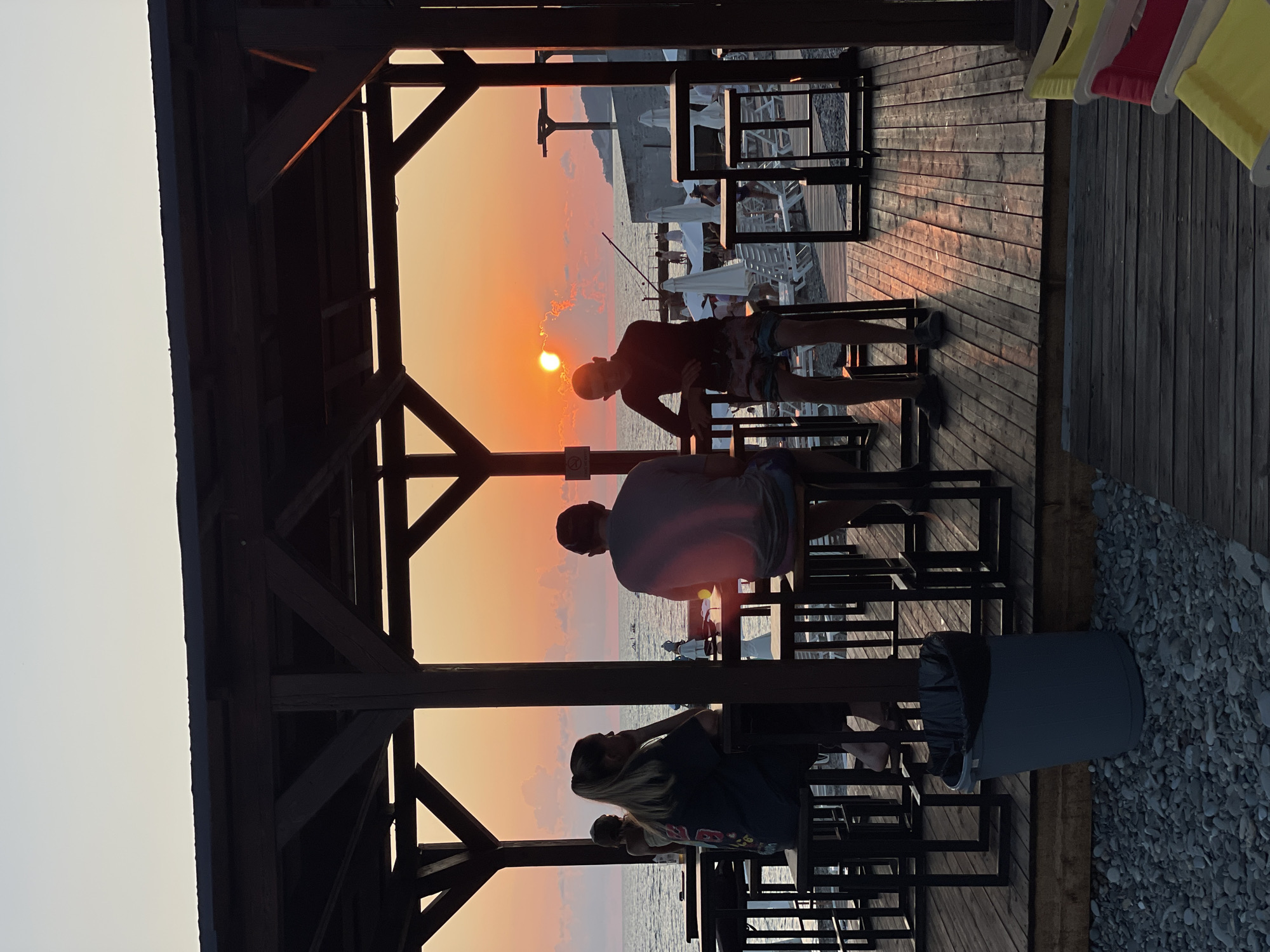 The image depicts a beachside bar at sunset. Three people are seated at tables under a wooden canopy. The sun is setting over the sea, casting a warm orange glow across the sky and reflecting on the water. In the foreground, a gray trash can rests on a pebbled surface. The wooden deck of the bar is visible, with chairs and tables set up. The background shows lounge chairs and umbrellas on the beach, with a few people walking near the water's edge. The location appears to be a resort area in Sochi, Russia, given the typical scenery of the Black Sea coast.