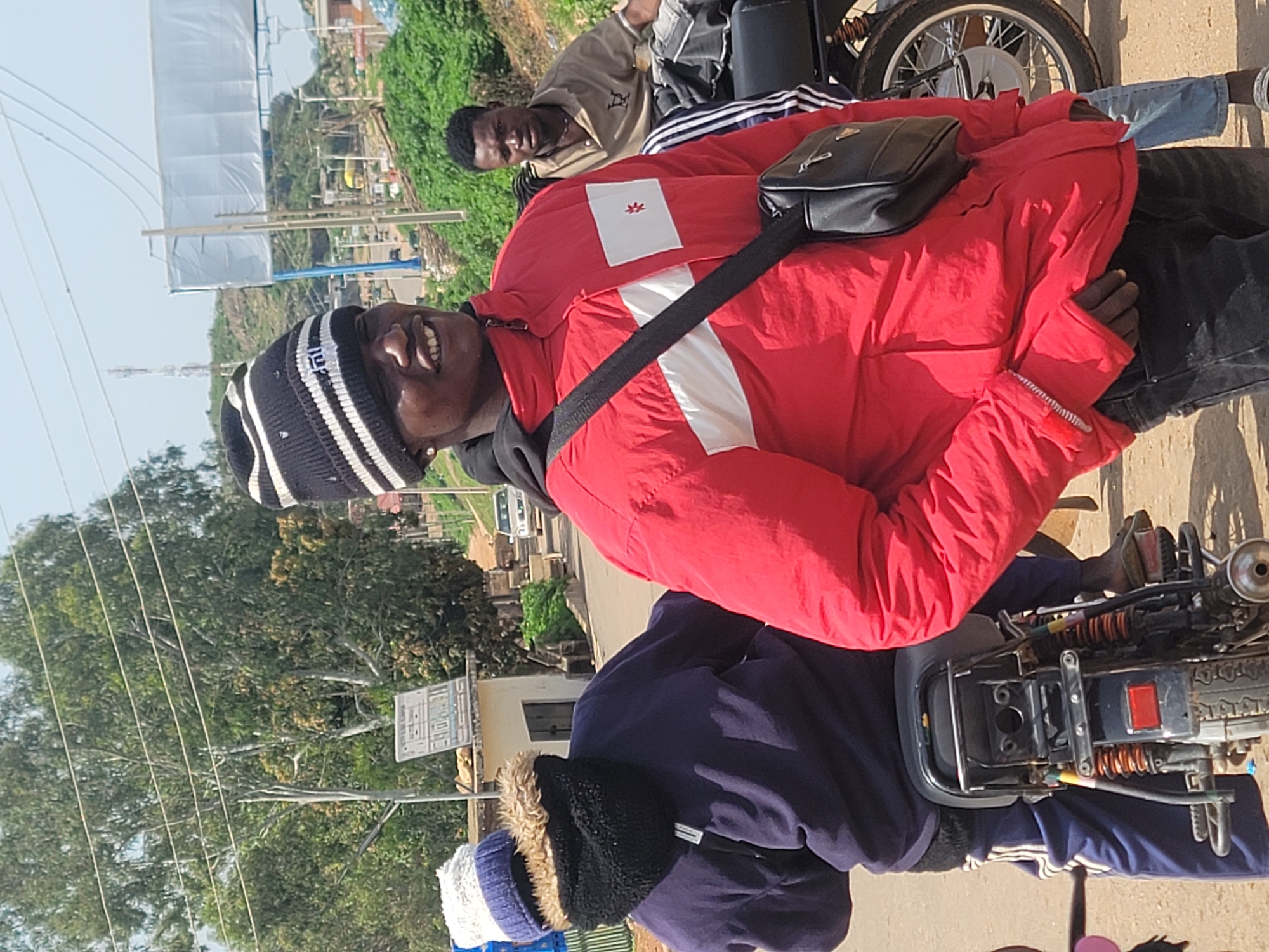 The image depicts a sunny day scene on an unpaved road, possibly in the city of Gura Top, Nigeria. In the foreground, a young Black man wearing a red jacket, a black and white striped beanie, and a black crossbody bag smiles at the camera. To his left, another person is partially visible, wearing a dark blue long-sleeved shirt, a blue and white beanie, and a black scarf around their neck. They are seated on a motorcycle, facing away from the camera. In the background, another Black man wearing a tan shirt is seated behind them on a motorcycle. The environment features lush green trees, a small building with a sign, and utility poles with wires. Further in the background, hills and a road with a few vehicles can be seen. The overall atmosphere suggests a casual outdoor gathering.