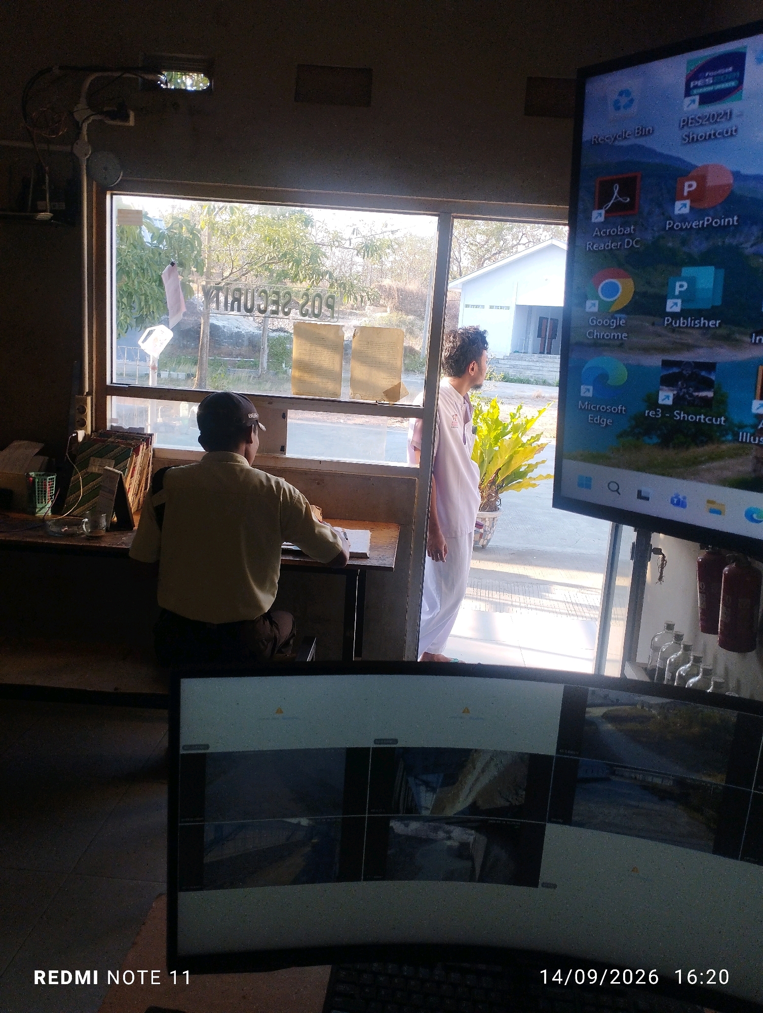 In Bero, Indonesia, a security guard sits at a desk in a booth, facing a window. The guard is wearing a light-colored uniform and a dark baseball cap. Outside the window, a white building and trees are visible. The booth is equipped with a computer monitor displaying security camera feeds. The monitor shows multiple camera views, including what appears to be an outdoor area with a road and a bridge. The scene suggests a security checkpoint or monitoring station.