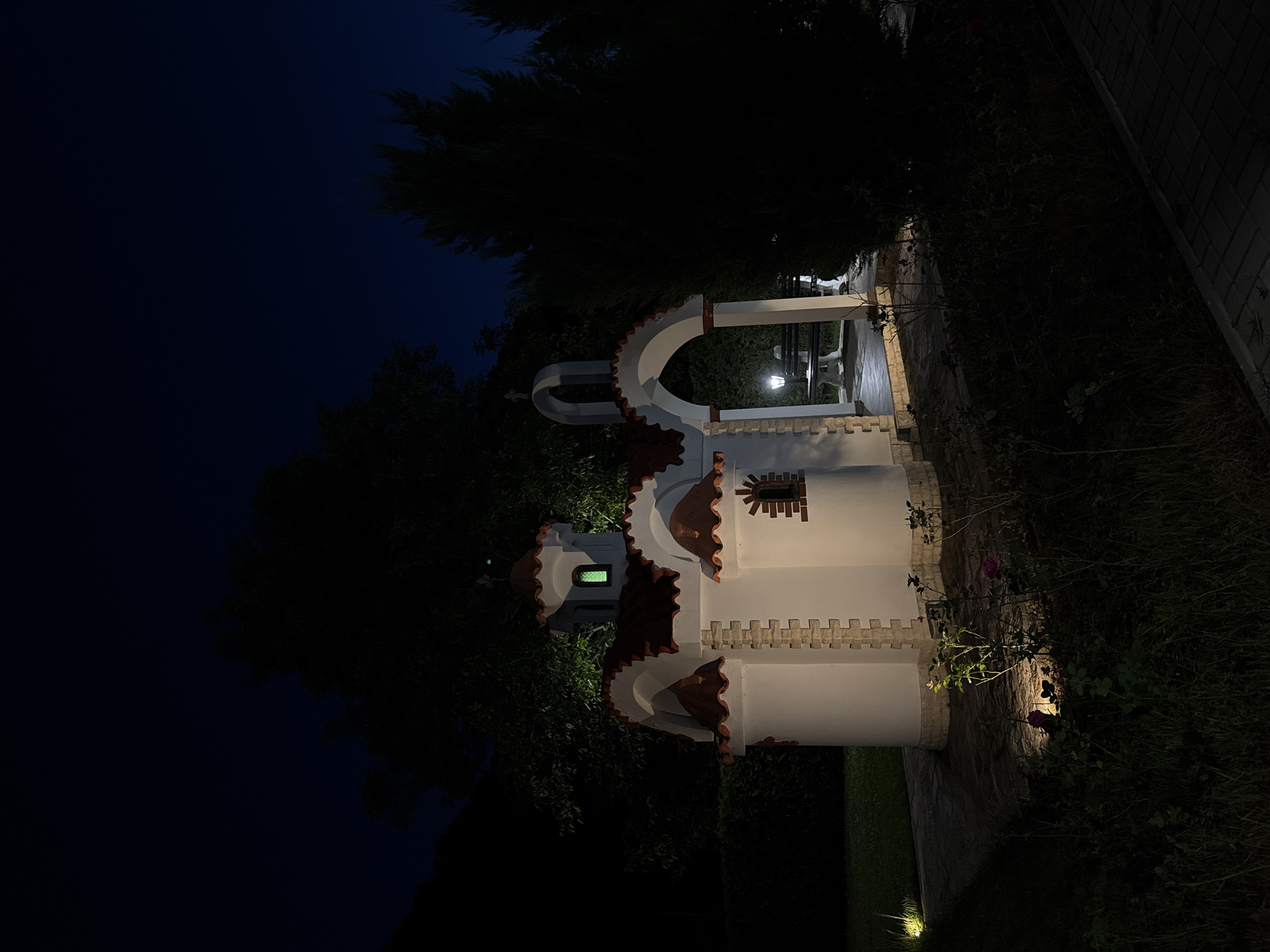 The image depicts a small, white, chapel-like structure with red-tiled roofs, illuminated by spotlights at night. The chapel features a central arched entrance, a small green-lit window, and decorative brickwork. It is situated in a garden setting with dark trees and foliage surrounding it. A paved walkway leads to the chapel, and there are benches visible under the archway. The scene is set in an outdoor environment with a dark blue night sky. There are no people visible in the image. The specific location in Gabrovo, Bulgaria is not identifiable from the image itself.