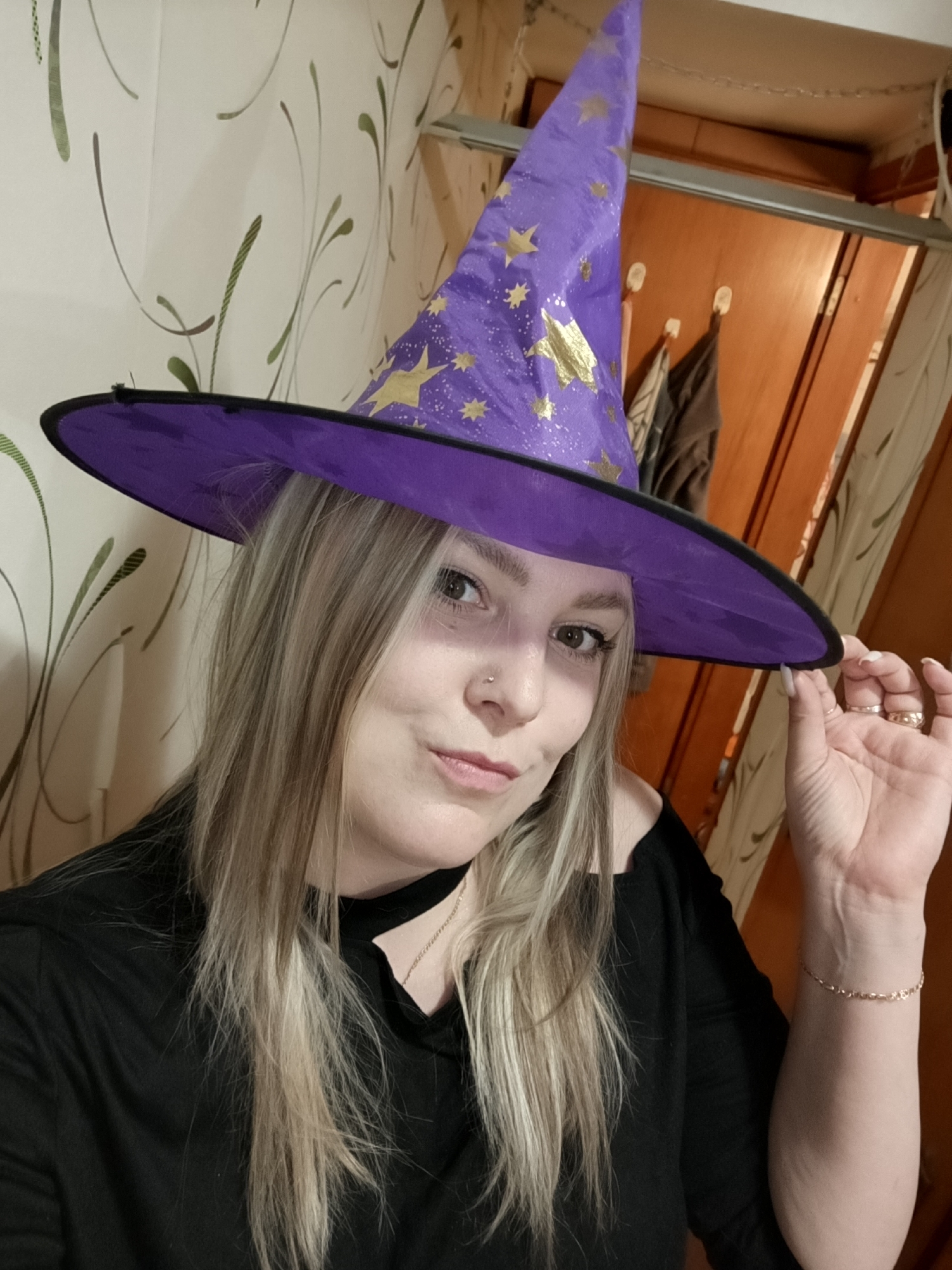 A young woman is captured in a head-and-shoulder selfie, holding the brim of a purple witch hat adorned with gold stars and glitter. She has long, straight blonde and brown streaked hair. Her makeup includes defined eyes and a light lip color, and she has a nose piercing. She wears a black long-sleeved top with a small cutout near the collarbone, a delicate gold chain necklace, a matching gold bracelet on her right wrist, and several rings on her right hand. Her fingernails are painted a light, neutral color.
She gazes directly at the camera with a soft, playful smile, subtly parting her lips. Her right hand gently holds the brim of her hat, contributing to her pose.
The background shows an indoor setting. To the left, there's a light-colored wall covered in wallpaper with a subtle green and brown stylized leaf or vine pattern. To the right, a wooden door or cabinet is visible, above which are two white plastic hooks holding dark clothing items. The lighting appears to be artificial, suggesting an indoor photograph taken possibly in the evening.
The purple witch hat strongly indicates a costume, likely for Halloween or a themed event. The overall mood conveyed is playful and festive. There is no visible text in the image. The location context is Марковский, Russia.