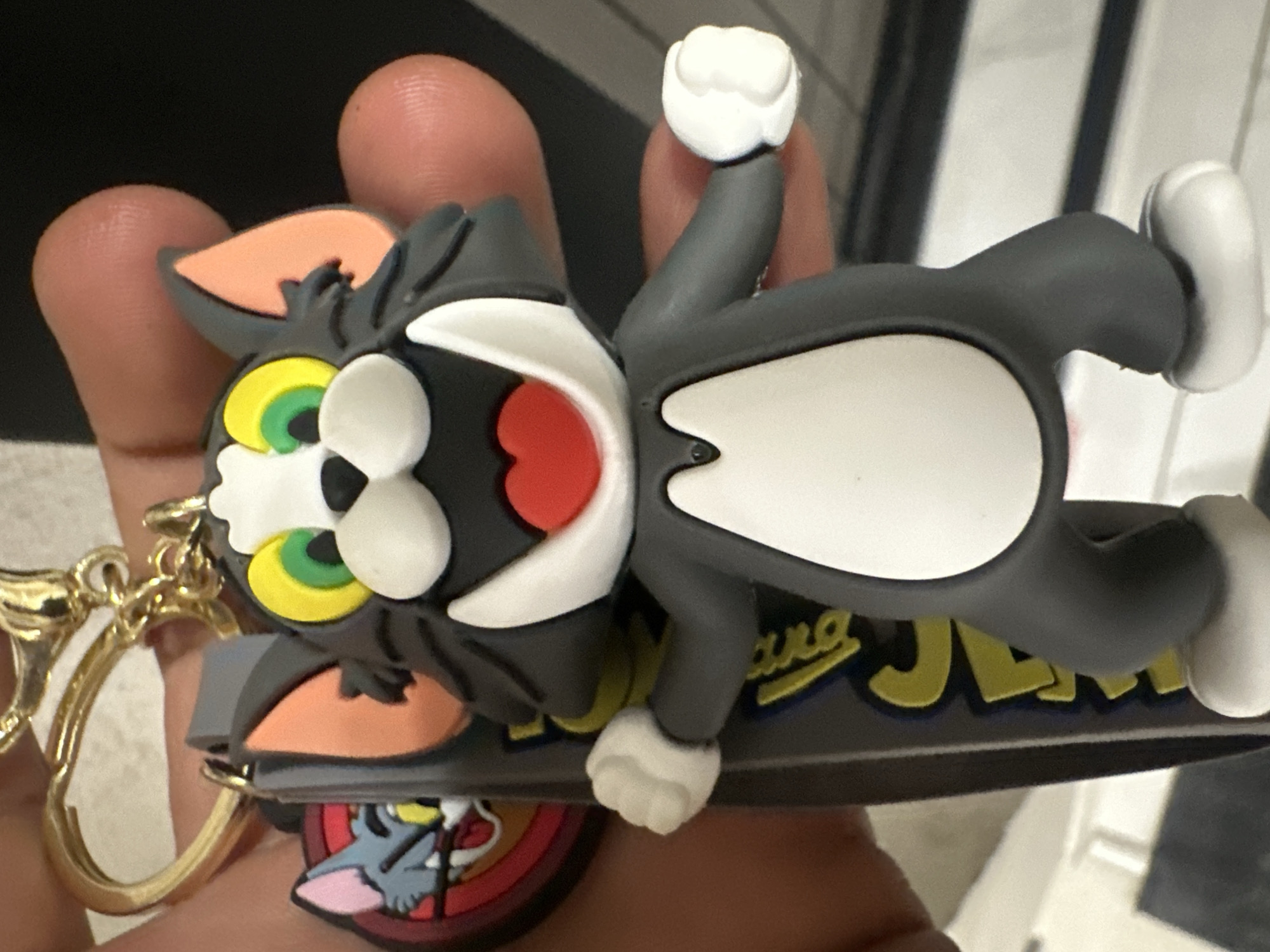The image depicts a multi-component keychain, featuring characters from the "Tom & Jerry" animated series, held by a human hand in Sofia, Bulgaria. The primary subject is a three-dimensional rubber or soft plastic figure of Tom Cat, approximately 7-8 centimeters in height, with a grey body, white face, chest, and paws. Tom's mouth is open, revealing a red tongue, and his eyes display yellow sclera with green irises. His inner ears are pink. He is depicted in an animated, standing pose with both arms slightly extended and fists clenched.
The Tom figure is attached at the top of its head to a gold-colored metal keyring. This keyring also holds a flat, dark grey strap, which features the yellow lettering "TOM & JERRY" in a cartoonish font. Additionally, a smaller, circular charm is attached, displaying a red background with a stylized depiction of Tom Cat's head, appearing to interact with a partially visible yellow element.
The keychain assembly is grasped by a human hand, with fingers and thumb visible on the left and right sides of the main Tom figure. The skin tone of the hand is light to medium. The background is dark and significantly out-of-focus, suggesting an indoor environment, with a faint lighter horizontal line potentially indicating a surface such as a countertop or table behind the hand. The scene is located in Sofia, Bulgaria.