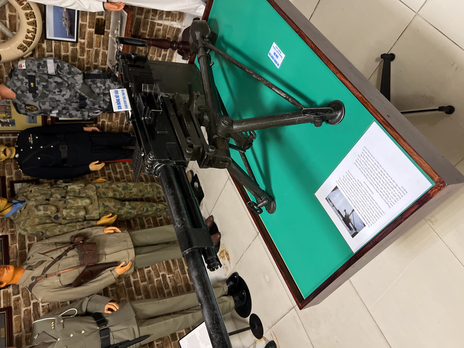 A collection of mannequins dressed in various military uniforms stands against a brick wall. A machine gun is mounted on a green-topped display table. The table includes explanatory text and an image depicting a similar gun. The angle of the photo provides a view down the barrel of the gun, with the mannequins in the background appearing small.