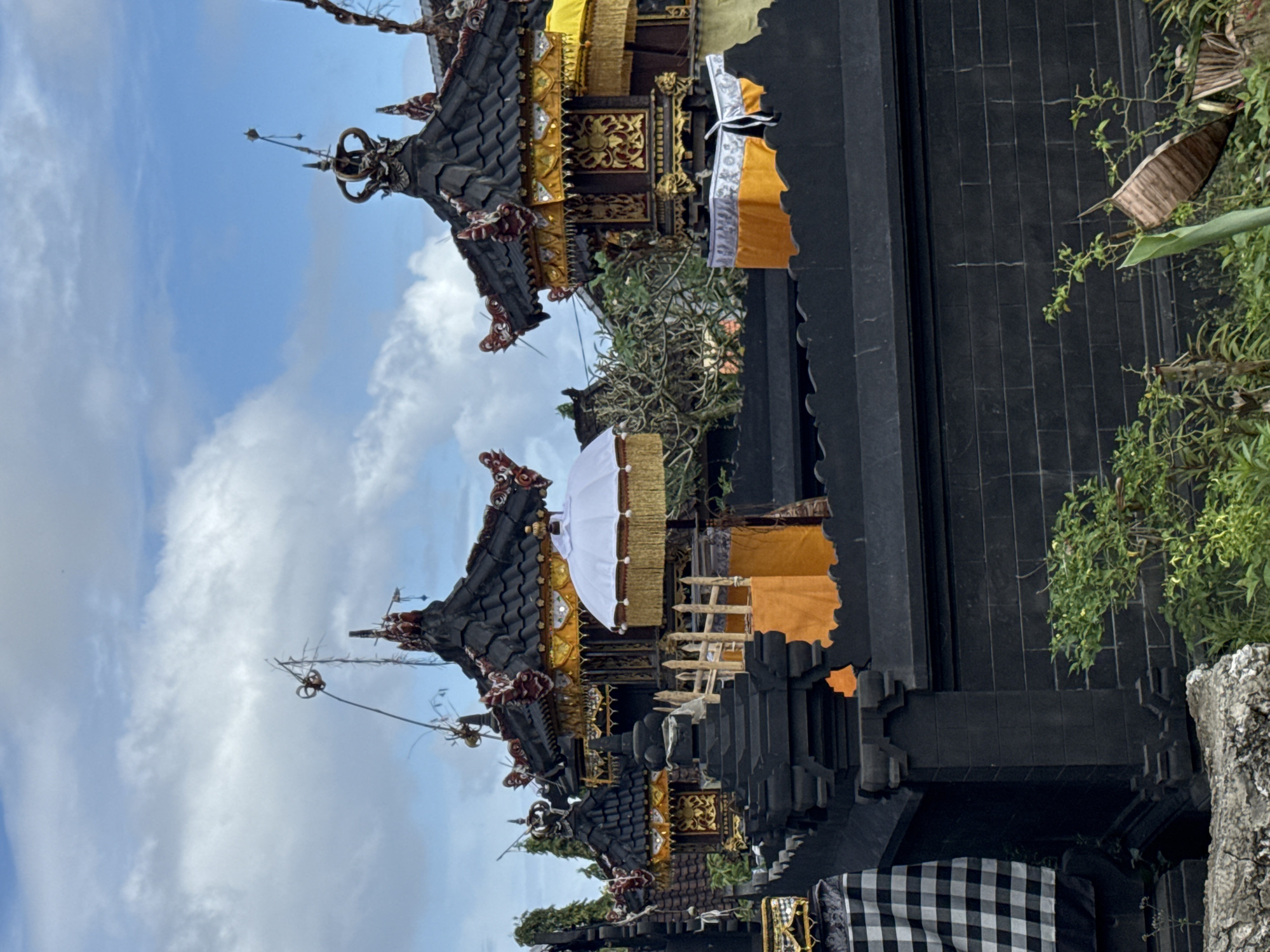 The image captures a scene from Kuta Selatan, Indonesia, featuring a temple with traditional Balinese architecture. The temple structures are predominantly dark in color, possibly constructed from stone or dark wood, and adorned with intricate carvings and golden decorative elements. Multi-tiered roofs, common in Balinese temples, are visible, topped with ornamental spires. A white parasol is positioned near one of the structures. Orange and yellow banners or cloths hang from the temple, adding color. The background reveals a sky with scattered clouds. Plants and greenery are present on the right side of the image. The photograph is taken at an upward angle, emphasizing the height of the temple structures against the sky.