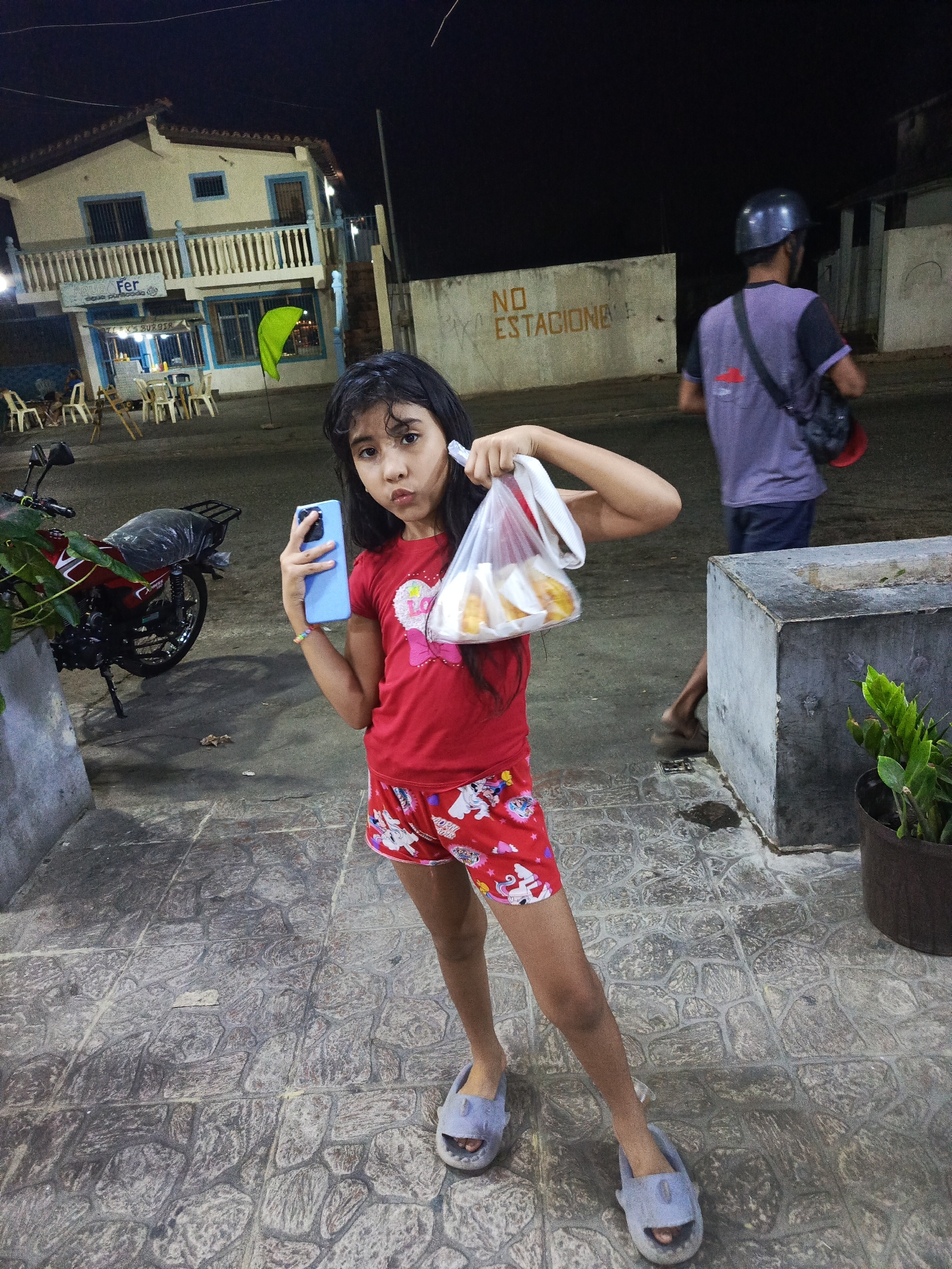 A young girl stands on a tiled street at night in what appears to be the city of Altagracia, Venezuela. She is wearing a red t-shirt, red shorts with unicorn patterns, and gray slippers. In her left hand, she holds a light blue smartphone. In her right hand, she holds a plastic bag containing what appears to be pastries or fried food. Behind her and to the left is a building with a business called "Aqua Fer." A red motorcycle is parked near the building. A man wearing a helmet, a gray shirt, and carrying a bag walks away from the camera to the right of the girl. In the background, there is a concrete wall with the words "NO ESTACIONE." A concrete planter with green plants is to the right of the girl. The street is paved with gray tiles.