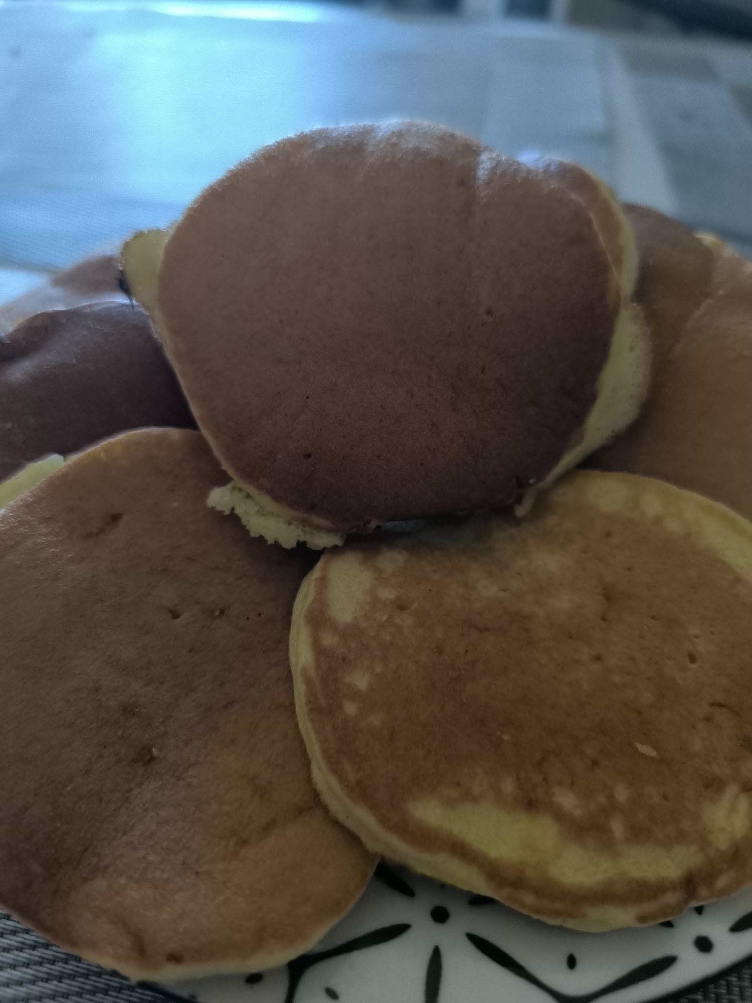 A close-up, overhead shot shows a stack of dorayaki on a decorative plate. The dorayaki are round, golden-brown pancakes with a lighter yellow interior. Some appear slightly darker than others, suggesting variations in cooking time or ingredients. The plate has a black and white floral pattern. The background is blurred, showing a light blue surface that could be a table or a window. The lighting is soft and diffused.