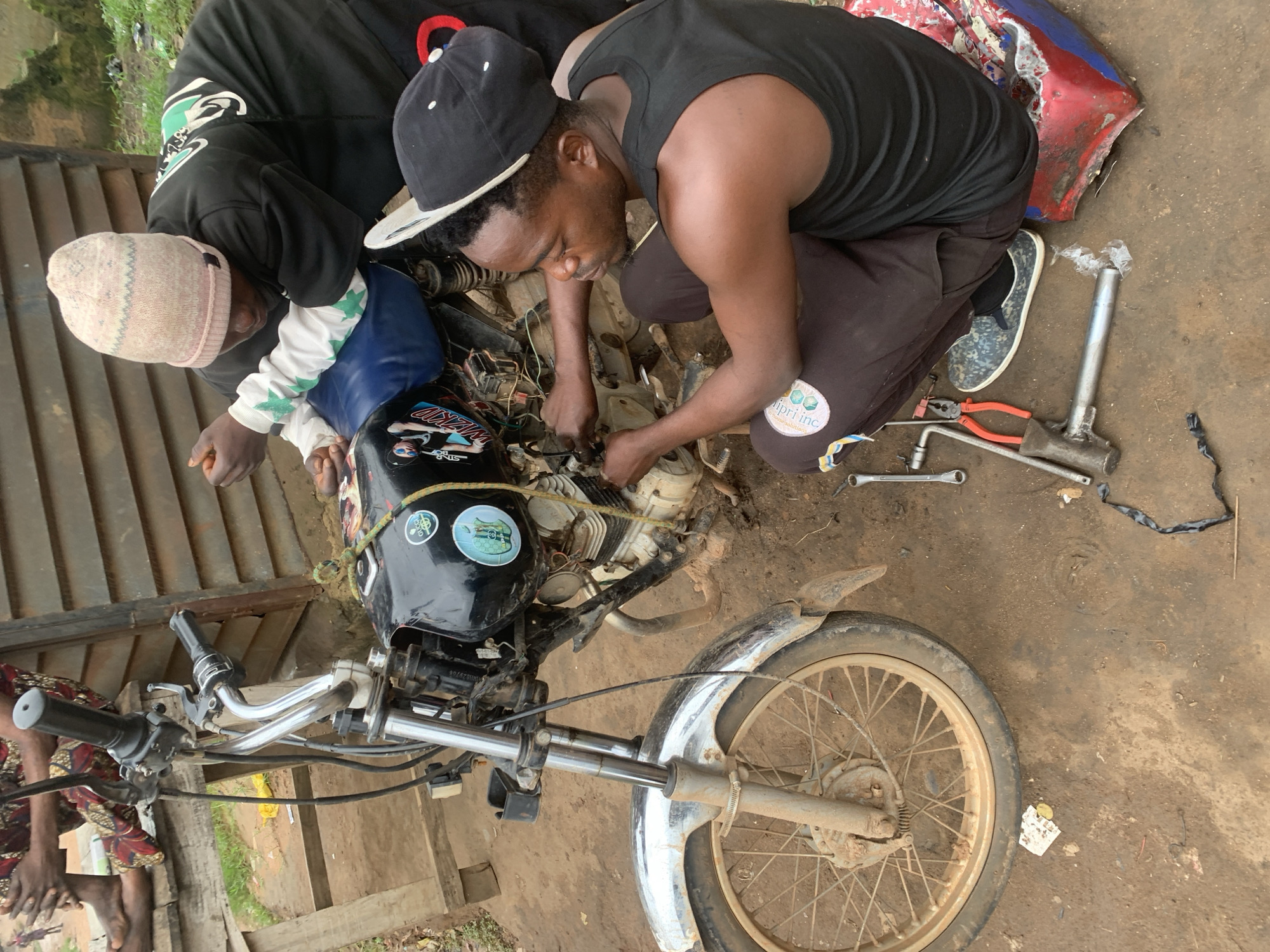 In the city of Shagamu, Nigeria, two individuals are observed working on a motorcycle. The scene appears to be an outdoor workshop or yard, with a dirt ground and a wooden structure in the background. One person, wearing a black tank top and dark pants with a patch, is crouched down and actively engaged with the motorcycle's engine, holding tools in their hands. This individual is wearing a black baseball cap. The second person, wearing a black long-sleeved shirt with white and green accents and a pink and white patterned beanie, leans over the motorcycle, also appearing to be involved in the repair. The motorcycle itself is black with blue accents on the seat and features some stickers. Various tools, including wrenches and pliers, are scattered around the workspace on the ground, along with a red and blue object that might be a container or part of the motorcycle. The overall impression is one of active mechanical work taking place in a somewhat informal setting.