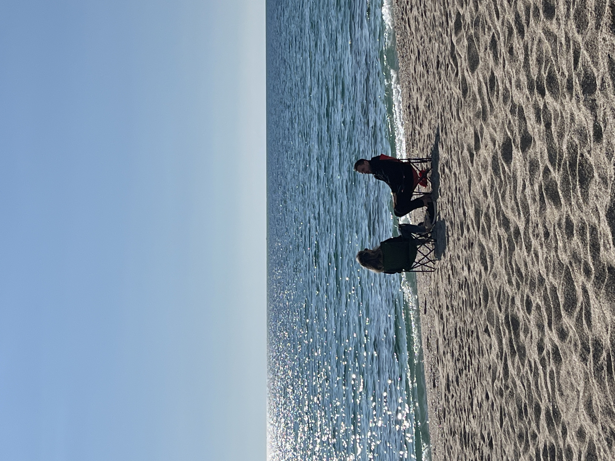 The image displays a sunny beach scene in Alanya, Turkey. A clear, light blue sky occupies the top portion of the frame. The sea, exhibiting a blue-green hue with numerous sun glints on its surface, stretches across the middle of the image, meeting the sky at a distant horizon. Small waves break gently along the shoreline. The foreground consists of light-colored sand, marked by ripples and depressions. Two individuals are seated on portable folding chairs on the sand near the water's edge. The individual on the left is seen from behind, characterized by long, light-colored hair and dark attire, seated in a dark-colored chair. The individual on the right, presenting as male, is viewed in left profile, wearing dark clothing, and positioned in a red folding chair. His head is turned slightly towards the left individual, and both are oriented towards the sea.