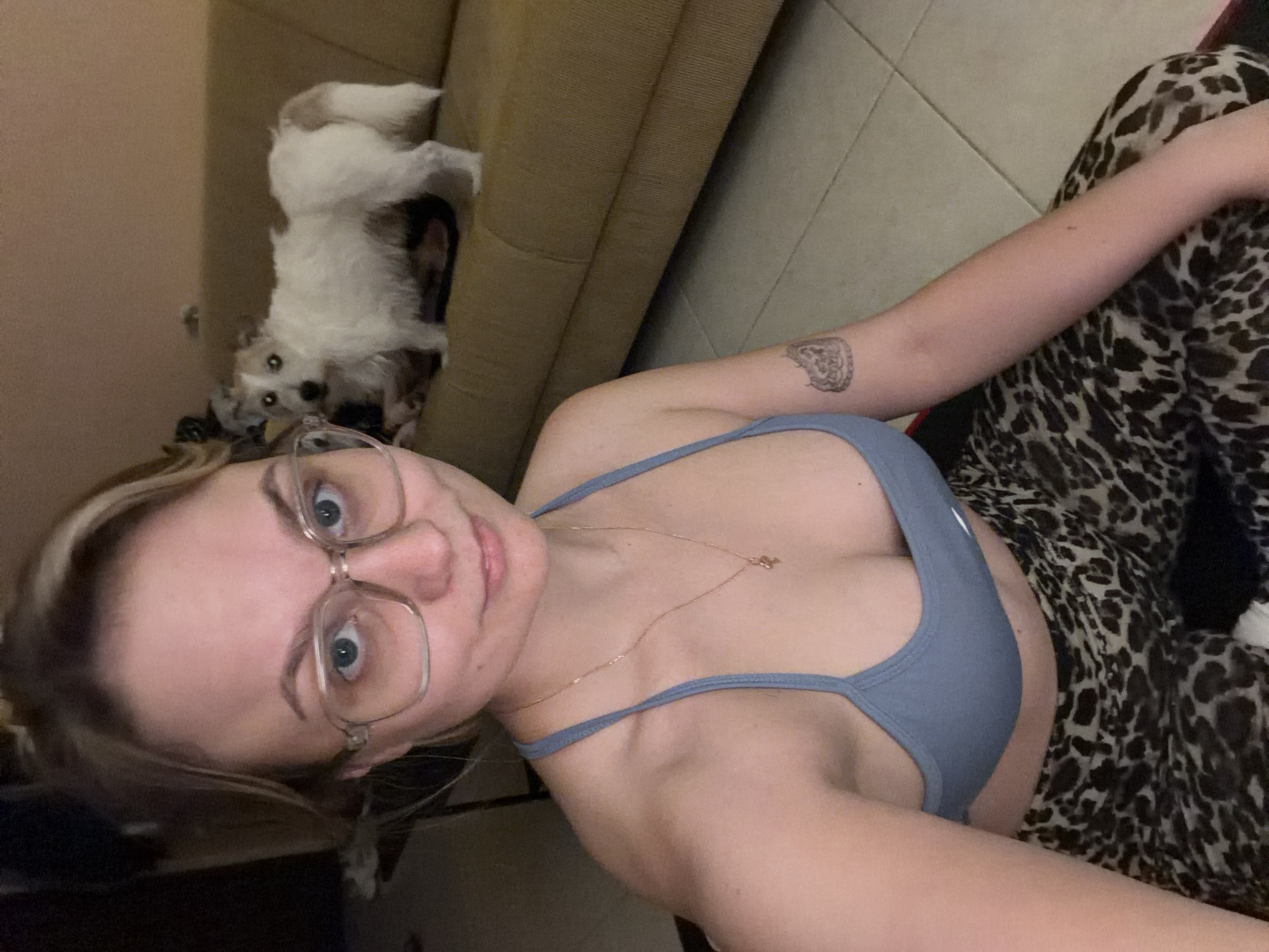 A woman wearing clear-framed glasses and a blue sports bra is taking a selfie indoors. She is lying down, with leopard-print pants visible on her right. A small white dog is visible on a beige couch in the background. It is not possible to determine if the photo was taken in Tel-Aviv, Israel.