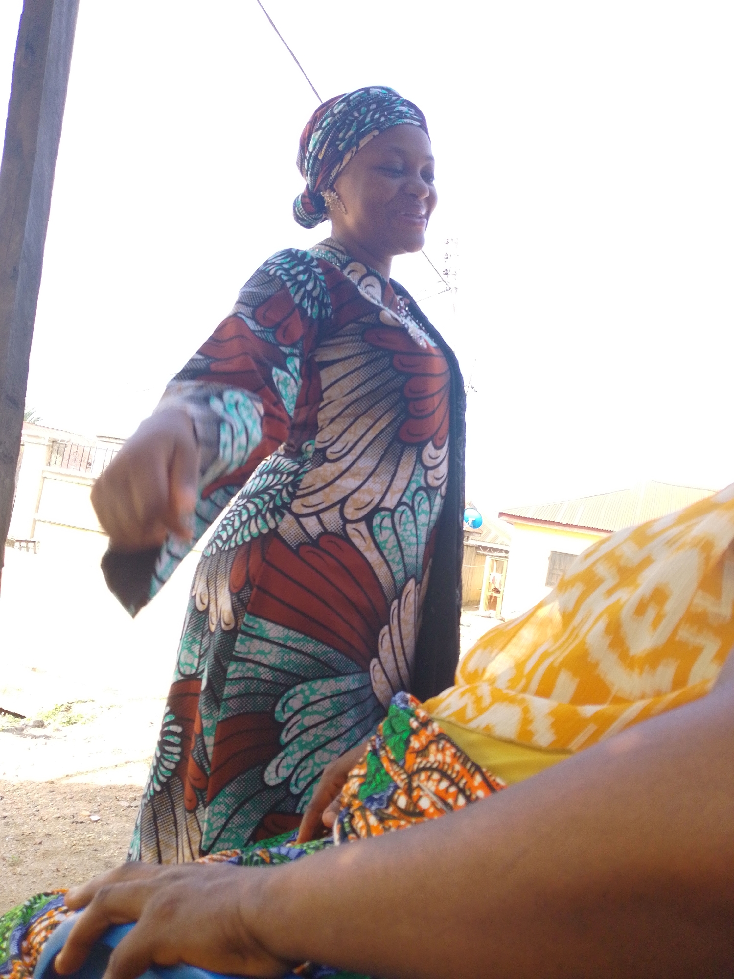 A medium shot depicts a woman in Lokoja, Nigeria, standing outdoors. She is wearing a long, colorful patterned dress and a matching headwrap. Her right arm is extended forward, with her hand slightly clenched. She is smiling and looking to her right. To her right, another person is partially visible, wearing a yellow patterned fabric. A portion of a wooden structure is visible on the left edge of the frame. In the background, there is a white building with a red roof and a blue object hanging from a wire. The ground appears to be dirt or gravel. The sky is bright, suggesting daylight.
