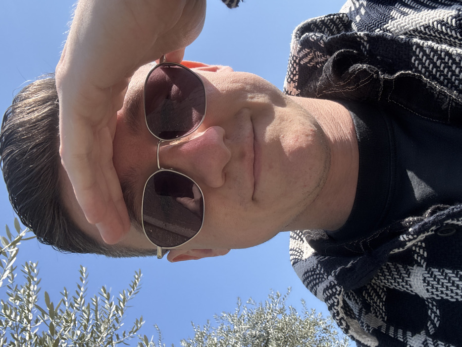 A close-up, upward-angled selfie depicts a young adult male outdoors in Peschiera del Garda, Italy, under a clear, bright blue sky. The male subject has short, dark brown hair and fair skin with light stubble visible on his chin and cheeks. He wears gold-framed, oval-shaped dark sunglasses and a black and white patterned woven jacket over a dark undershirt. His right hand is raised to his forehead, palm flat and fingers extended, shielding his eyes from direct sunlight. A subtle smile is present on his face. Reflections of a smartphone, presumably held by the subject to take the photo, are observable in both sunglass lenses. A segment of an olive tree with light green foliage is present in the soft-focus left background.