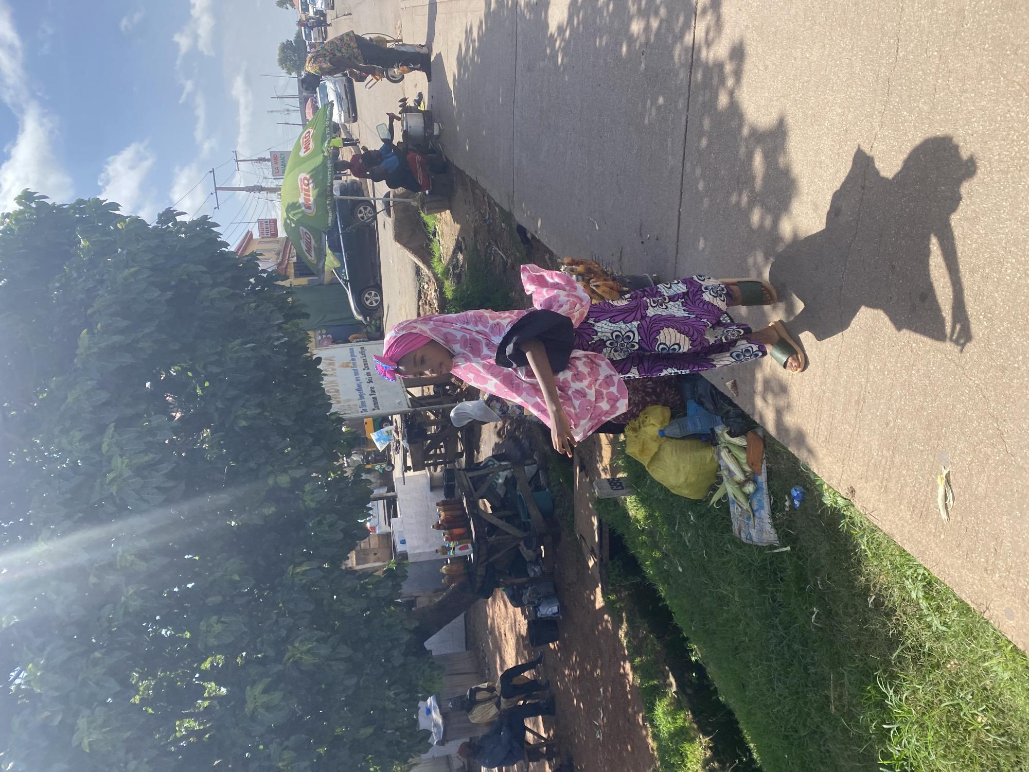 This media file depicts a street scene in Gura Top, Nigeria. The primary subject is a young woman wearing a pink and white patterned headscarf and a purple and white patterned dress, standing on a sidewalk. She appears to be a vendor, with a selection of goods laid out on the ground around her, including what looks like corn. Behind her, a wooden stall holds several bottles of liquids, possibly fuel or beverages. Further back, there are other people and vehicles, including a motorcycle and cars, suggesting a busy commercial area. A green umbrella with the MILO logo is prominently displayed, along with signs for "LA MED PHARMACY". The sun is shining, casting shadows on the ground, indicating it is daytime, likely morning or afternoon. The overall atmosphere seems to be one of everyday commerce and daily life in a Nigerian community. The visible text includes brand names like MILO and a pharmacy name, as well as a phrase that may be in Hausa: "To live together, we must live in peace Zaman Tare Sai do Zaman Lofiya".