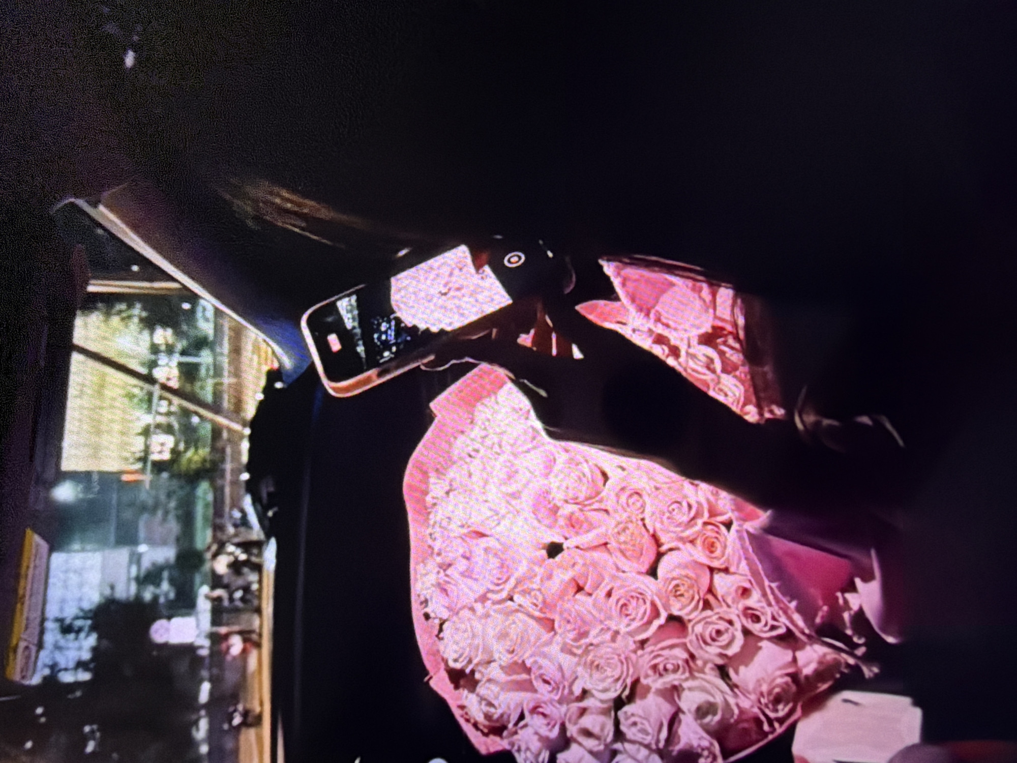 In a dimly lit, indoor setting, possibly a vehicle at night, a person's hand holds a smartphone to capture an image of a large bouquet of pink roses. The bouquet is the prominent subject, filling the lower left portion of the frame with its vibrant color and textured petals. The smartphone screen displays a bright, pixelated image, indicating it is actively being used for photography. Through the background, a blurred urban scene is visible, suggesting the presence of city lights and structures, hinting at a location within a city such as Beșalma, Moldova, although specific landmarks are not discernible. The overall atmosphere is one of intimacy and captured moment.