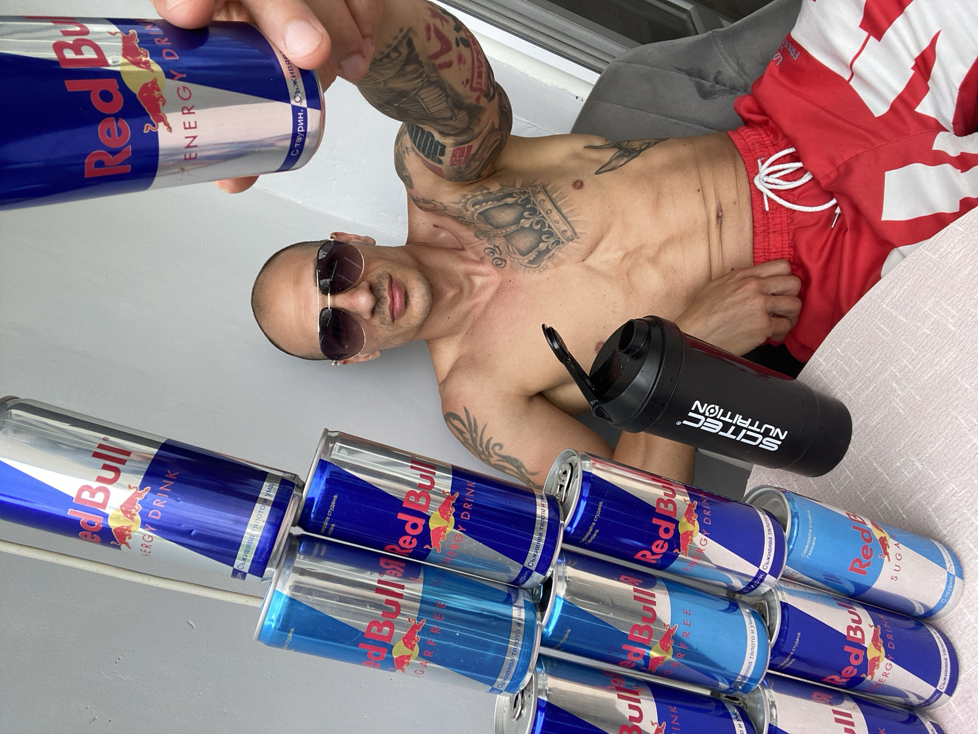 A man with a shaved head, sunglasses, and extensive tattoos on his chest and arms is holding a can of Red Bull. He is wearing red shorts with white accents. In front of him, a tower of Red Bull cans is stacked precariously, with a black shaker bottle resting beside them. The scene appears to be outdoors, possibly on a balcony or patio, with a light-colored wall in the background. The lighting suggests it is daytime. There is no indication of the location being in the city of Pleven, Bulgaria.