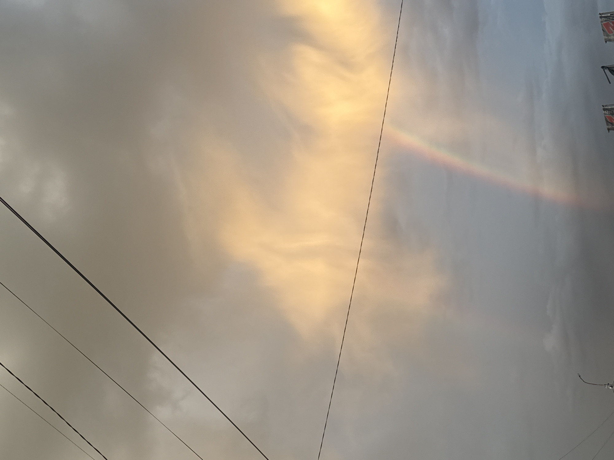 A rainbow arches across a cloudy sky in an urban setting. The sky is a mix of grays and whites, with a prominent band of warm, golden light illuminating some of the clouds in the upper portion of the image. Three dark power lines are strung diagonally across the sky, adding a geometric element to the natural scene. In the lower portion of the image, silhouetted against the dim sky, is a street lamp on the left, and three flags, two of which are partially visible on the right. The flags have red and white patterns. The overall impression is one of a dramatic sky, possibly after a rain shower, with the urban infrastructure in the background.