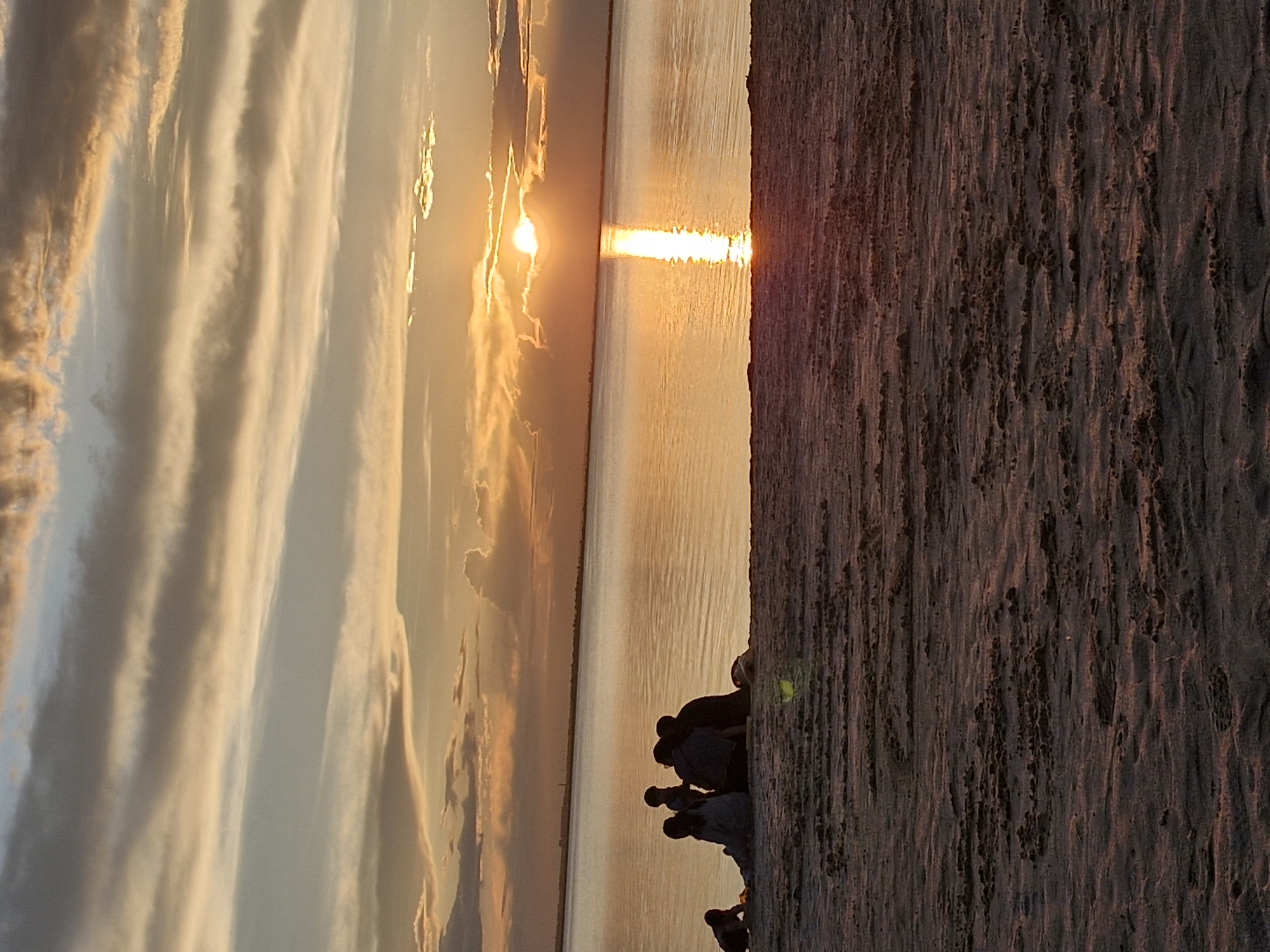 The image depicts a group of silhouetted figures gathered on a sandy beach, facing a body of water. The sun is setting over the water, casting a warm, golden glow and a reflection on the surface. The sky is filled with dramatic clouds, streaked with shades of orange, yellow, and grey. The foreground is the textured sand of the beach. Based on the provided information, this scene is located in Kokshetau, Kazakhstan. The figures appear to be a family or friends enjoying the sunset. There are no specific landmarks or city elements visible in the image that would allow for further localization within Kokshetau.