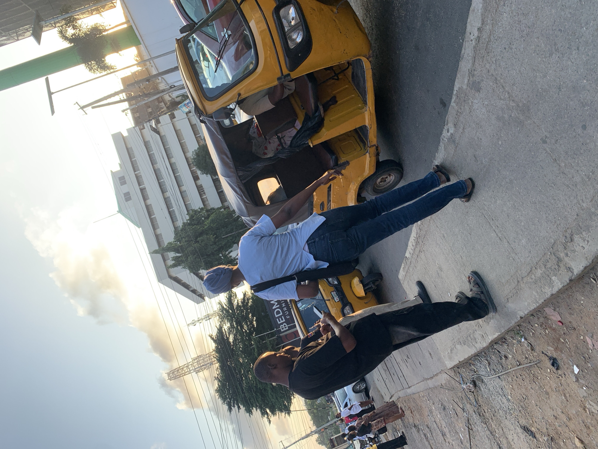 A group of people are standing on a sidewalk next to a street in Ikeja, Nigeria. The scene is bathed in the warm light of a setting sun, casting long shadows. A yellow auto-rickshaw, also known as a "Keke," is parked on the right side of the street. Two men are in the foreground, one in a black t-shirt and dark pants, looking down at his phone, and the other in a light blue t-shirt and jeans, facing away from the camera. A woman wearing a headscarf is also visible in the auto-rickshaw. In the background, there are buildings, trees, and power lines, suggesting an urban environment. Several other people and vehicles are present on the sidewalk and street, indicating a busy area.