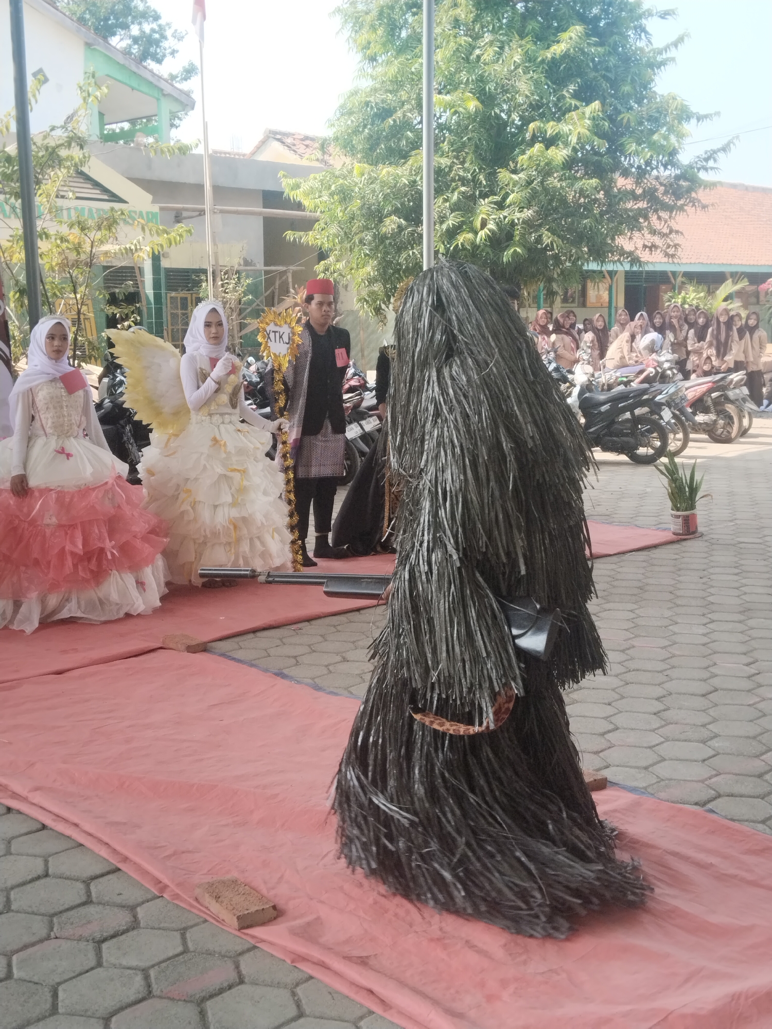 On a paved area in front of a building, possibly a school in Pakulaut, Indonesia, a group of people are gathered. In the foreground, a person in a dark, shaggy costume stands on a pink tarp. To the left, two individuals in elaborate dresses and headscarves are positioned on the same tarp. One wears a white dress with yellow wings, while the other has a pink and white dress. Behind them, a man in traditional attire with a red cap holds a decorative sign. A crowd of onlookers, many of whom appear to be students in uniforms, are visible in the background, along with several parked motorcycles. The scene suggests an outdoor event or performance.