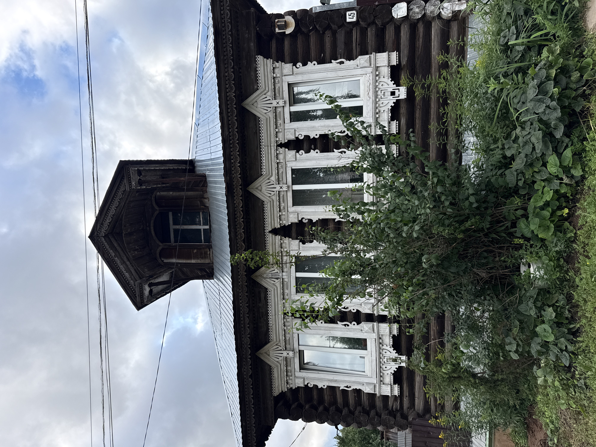 This is a medium shot, eye-level, outdoor, full-color photograph of a traditional wooden house in Votkinsk, Russia. The house is constructed from dark, weathered logs. It features an ornate, white wooden trim around its windows, with intricate carvings. The windows themselves are multi-paned and reflective, showing the sky. A metal roof is visible above the window level, and a small, dark wooden dormer with a pointed roof juts out from the main roofline. The house is partially obscured by lush, green foliage and trees in the foreground. The sky is overcast with patches of blue peeking through. A sign with the number "45" is visible on the right side of the house. Electrical wires are strung across the upper portion of the frame. No people are present in the image.