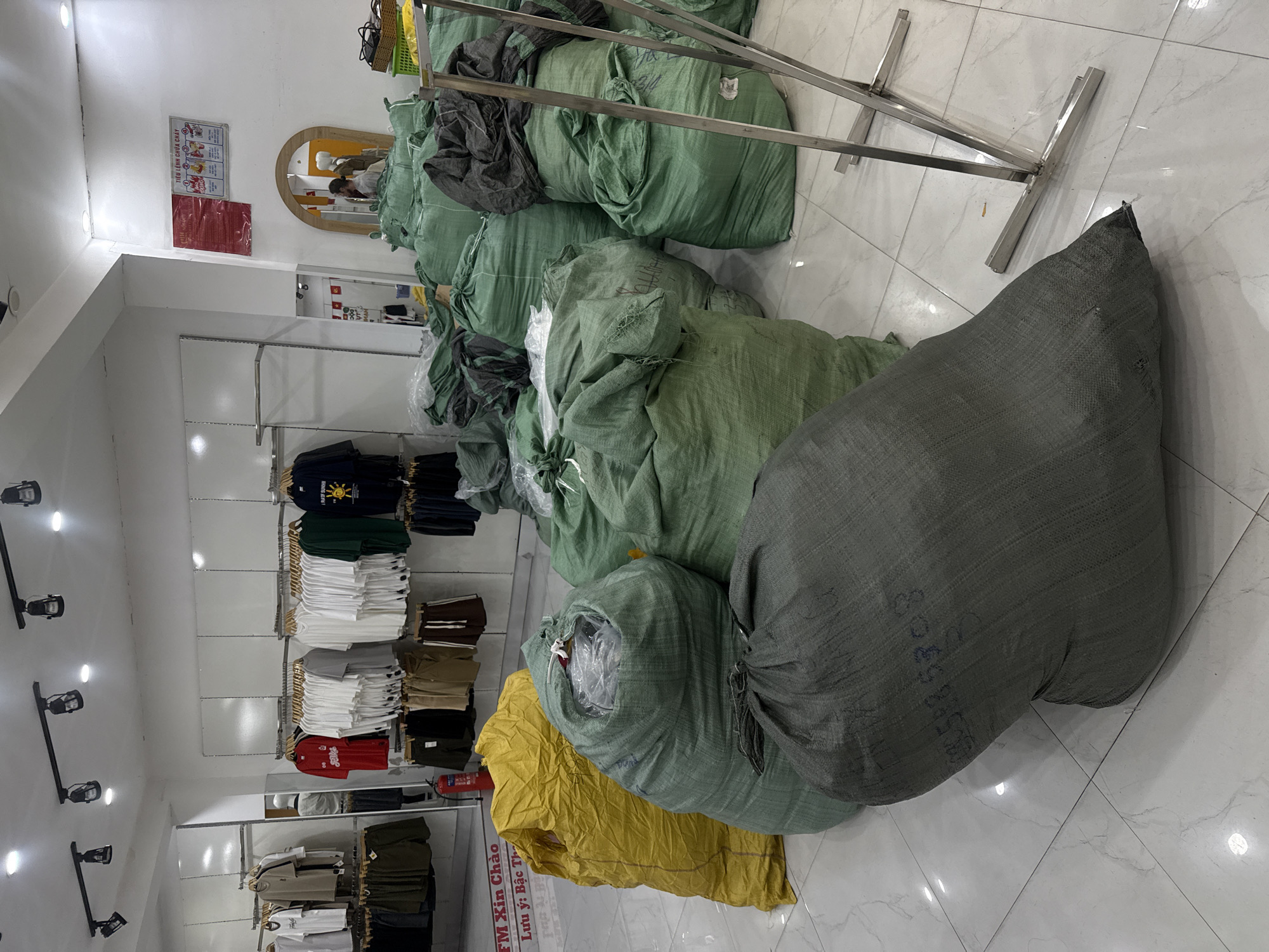 The image shows the interior of a clothing store in Da Lat, Vietnam. The store is filled with large, green, burlap sacks, likely containing clothing or textiles, piled on the floor. In the background, clothing racks display various t-shirts and shorts. There is a fire extinguisher mounted on a wall. A sign in Vietnamese is visible on the wall near the entrance, along with a mirror. A person is partially visible in the mirror, appearing to be a shop assistant. The lighting suggests it is daytime.