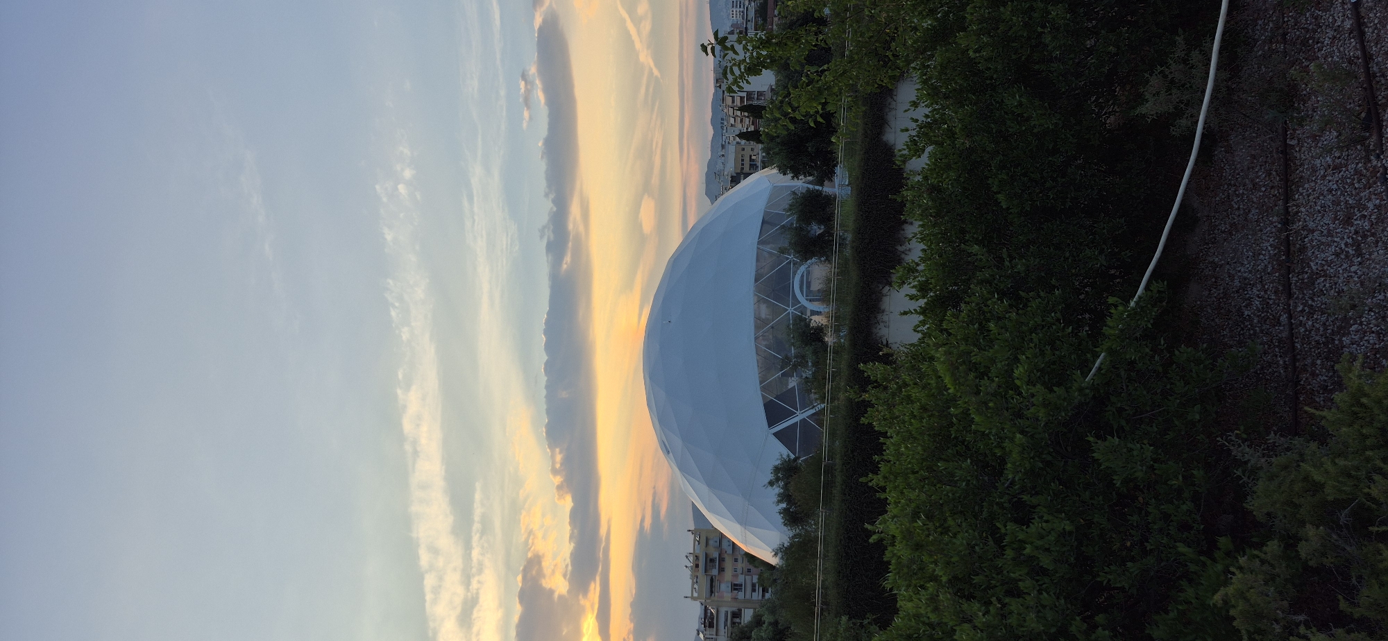 The image captures a geodesic dome structure, possibly a sports arena or event venue, bathed in the warm light of a sunset. The dome is white and has a distinct geometric pattern of triangular panels. It is situated in Kallithea, Greece, with buildings and trees visible in the background, suggesting an urban or suburban setting. The foreground is filled with dense green foliage, obscuring the lower portion of the dome and creating a natural barrier. The sky above is a soft gradient of blues and oranges, with wispy clouds reflecting the sunset's hues. There are no people visible in the frame.