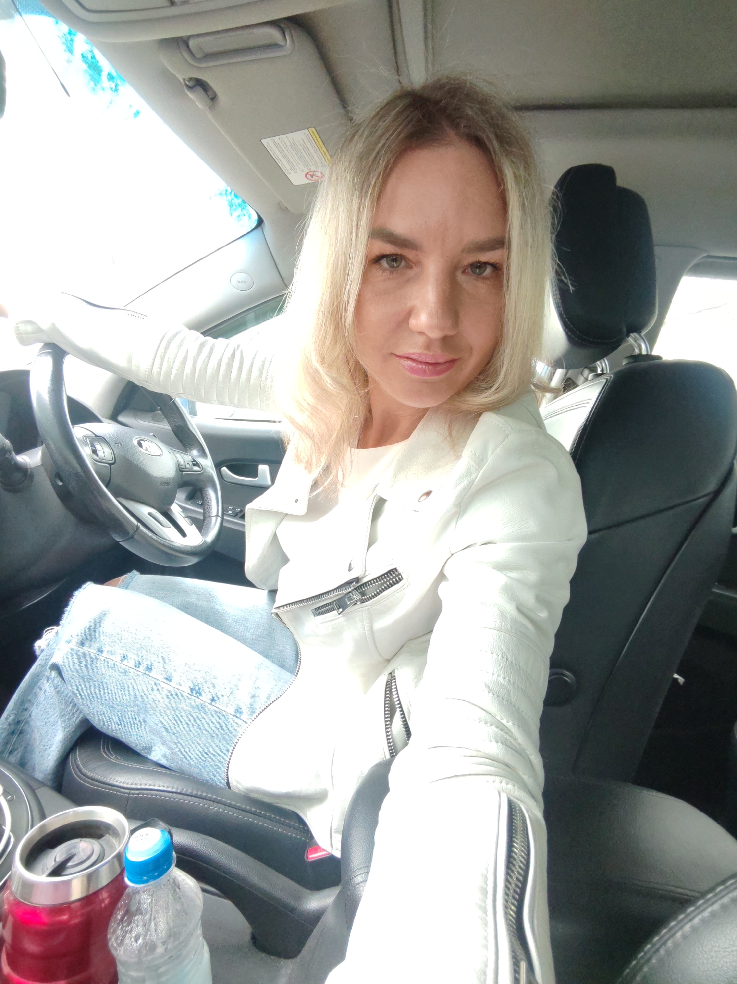 In a bright, sunlit car interior, a blonde woman in a white leather jacket and distressed denim jeans sits in the driver's seat, holding the steering wheel with her left arm. Her right arm is bent, with her hand extended towards the camera as if taking a selfie. A red thermos and a water bottle are visible in the center console. The car appears to be stationary, with the bright sunlight suggesting it is daytime. The environment is not identifiable as being in a specific city or location.