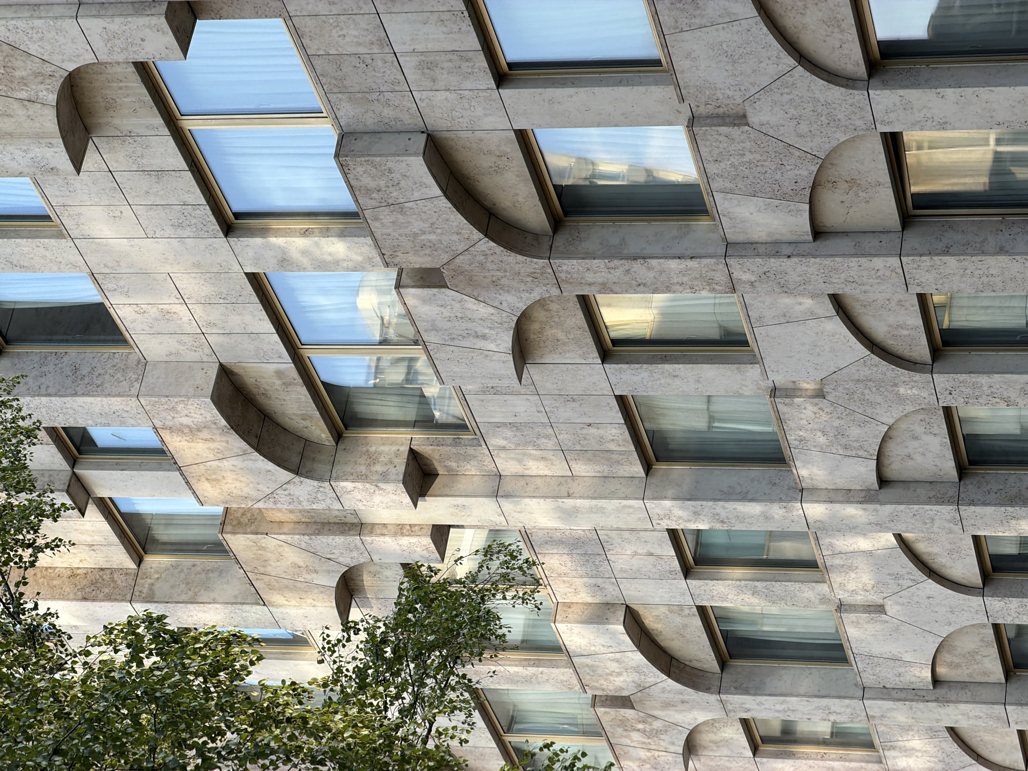 The image shows a close-up of a building's facade in Luxembourg City, Luxembourg. The building is constructed with light-colored stone tiles and features a repeating pattern of arched recesses and rectangular windows. Sunlight illuminates the textured stone, casting subtle shadows that highlight the architectural details. A portion of a leafy green tree is visible in the upper left corner, its branches extending towards the building. The windows have golden frames and are covered with sheer white curtains. The overall impression is one of modern architecture with classical influences.
