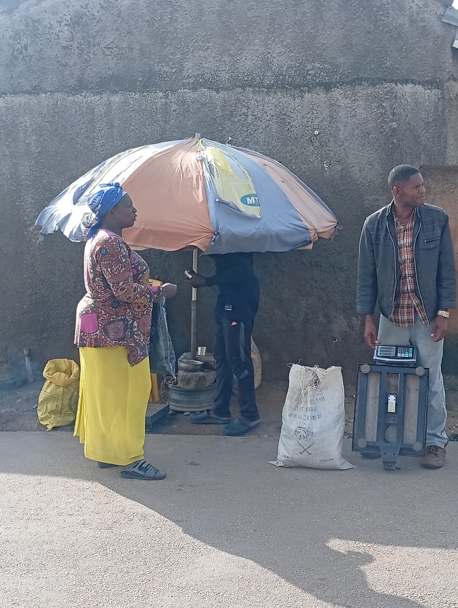 In Jos, Nigeria, two individuals are engaged in a transaction beneath a large, colorful umbrella. A woman, adorned in a vibrant patterned top and a flowing yellow skirt, stands facing left. To her right, a man in a grey jacket over a plaid shirt stands behind a digital scale. Between them, a white sack with Chinese writing and graphics is placed on the ground. In the background, a rough-textured concrete wall forms the backdrop, with a few yellow sacks visible to the left. The scene is set on an asphalt surface, likely a street or market.