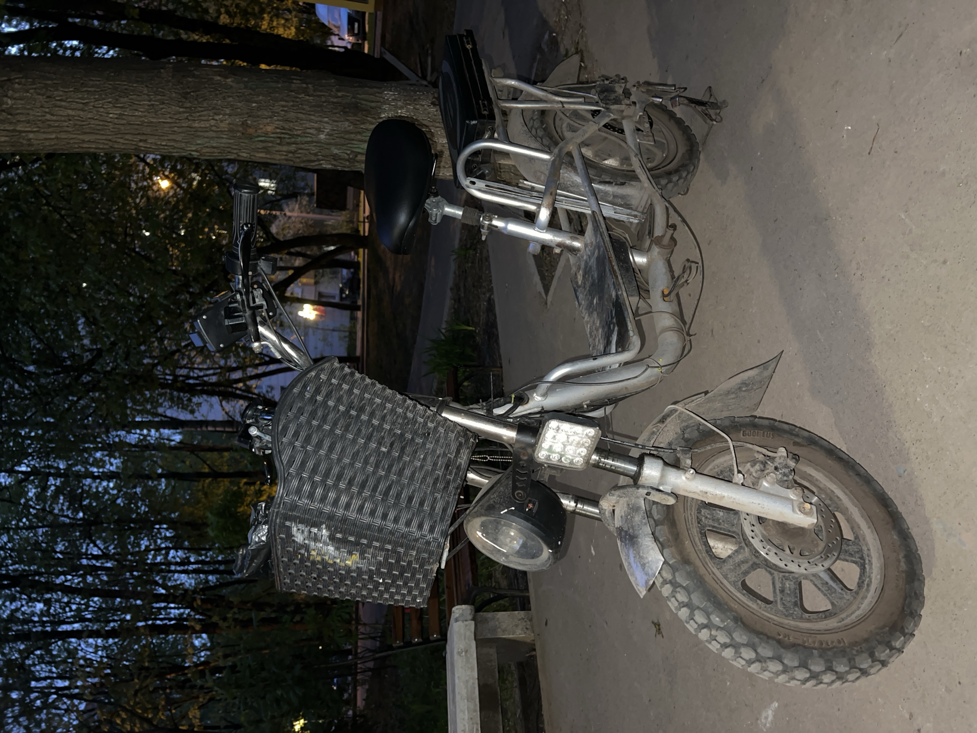 The image shows an electric bike parked outdoors. It has a silver frame, black seat, and a large woven basket on the front. The bike has lights mounted on the handlebars and a rear case behind the seat. The bike is parked on concrete or paved ground. The background includes trees and a building, suggesting it may be located in a park or residential area. The image was taken in Khimki, Russia.