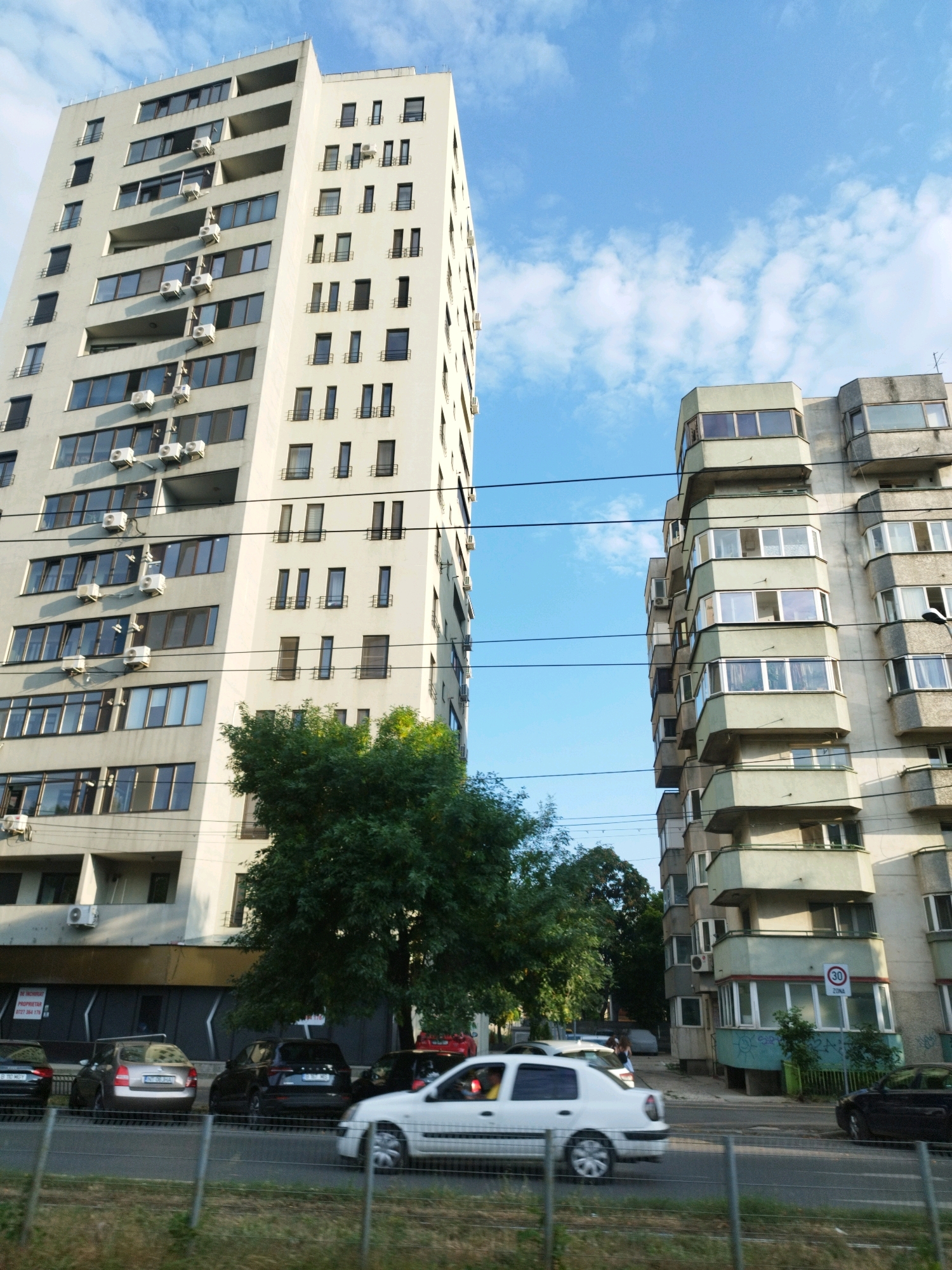 The image depicts a street scene in Bucharest, Romania, featuring two tall residential buildings. The building on the left is a modern, white, multi-story apartment complex with numerous balconies and windows. The building on the right is an older, grey, multi-story structure with distinctive protruding balconies, some of which are enclosed with windows. The sky above is blue with scattered white clouds. In the foreground, a street is visible with several cars, including a white sedan, a dark SUV, and other vehicles. Trees and greenery are present between the buildings and along the street, suggesting an urban setting with some vegetation. Electrical wires are strung across the scene, typical of city infrastructure. One of the buildings has a speed limit sign displaying "30 ZONA". The overall impression is a view of a typical urban street in Bucharest on a clear day.