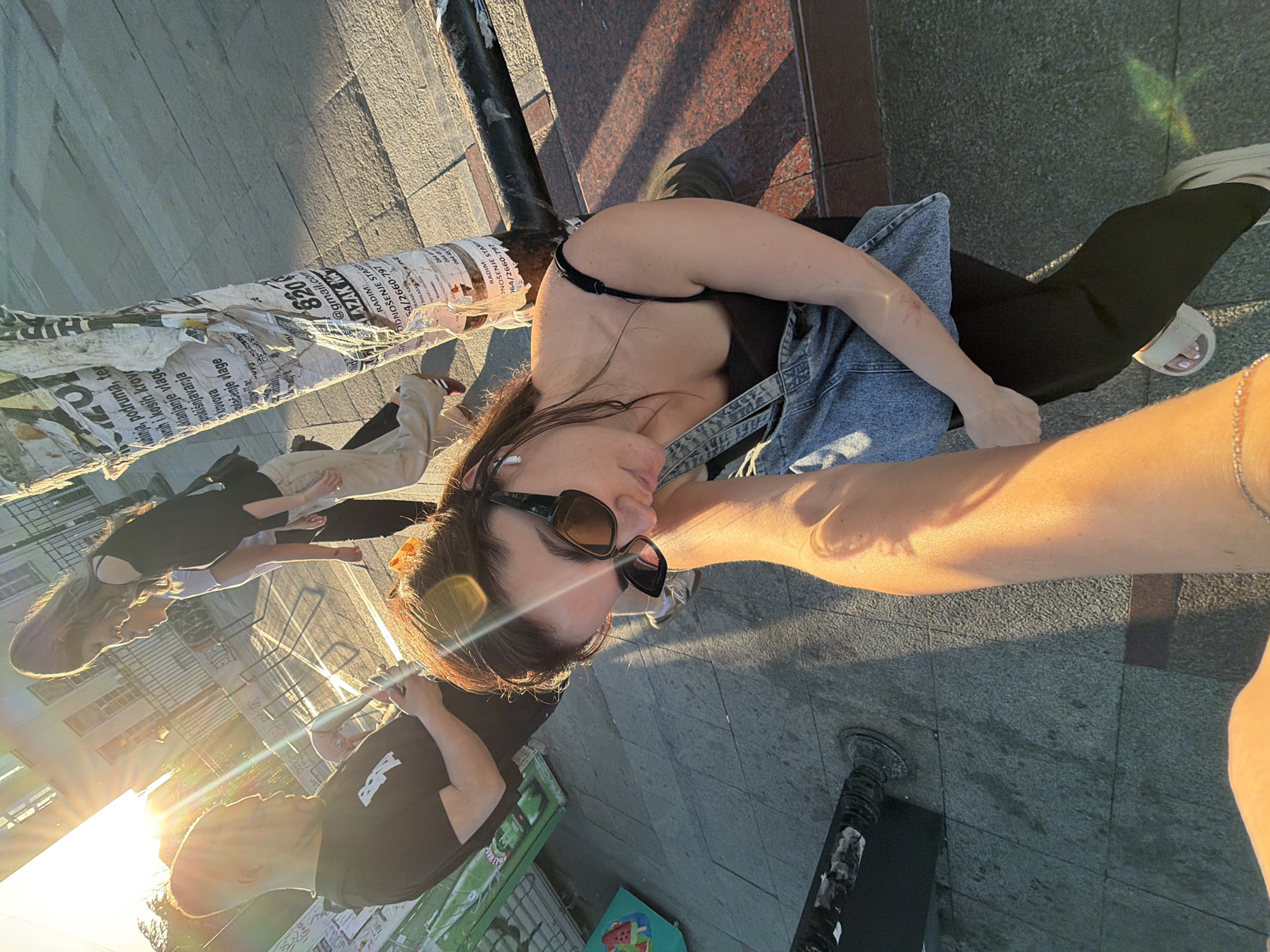 A high-angle selfie perspective shows a woman with sunglasses, wearing a black top and denim overalls, standing on a city street in Palilula Urban Municipality, Serbia. Sunlight illuminates her and the surroundings. The background features other people, including a person in a black t-shirt, and urban elements like a trash can and a pole covered in posters with discernible text. The ground is paved with gray stone, and a portion of a building is visible in the reflection.