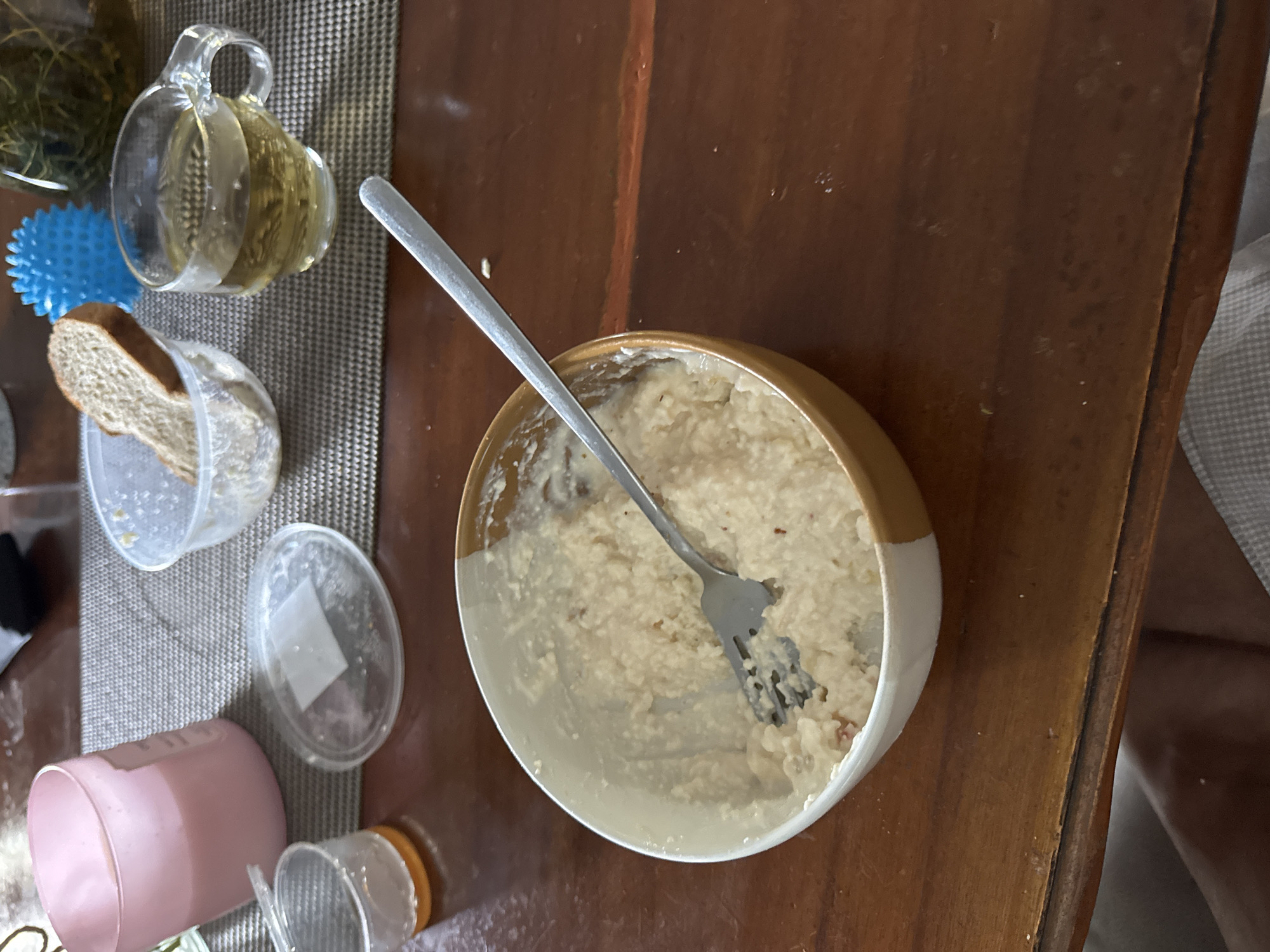 The image depicts a close-up, overhead view of a wooden table set with breakfast items. In the foreground, a bowl of oatmeal with a fork resting inside is centrally placed. To the left, a pink candle in a glass holder and a small plastic cup with an orange liquid are visible. In the background, a clear glass mug filled with amber liquid, likely tea, sits next to a clear plastic container holding a slice of bread. A blue spiky ball and a glass jar with green content are also in the background. The table is covered with a textured placemat. The setting appears to be an indoor environment, possibly a dining area. There are no people visible in the image. The lighting suggests it could be daytime. The image does not provide information about its location in Kuta Selatan, Indonesia.