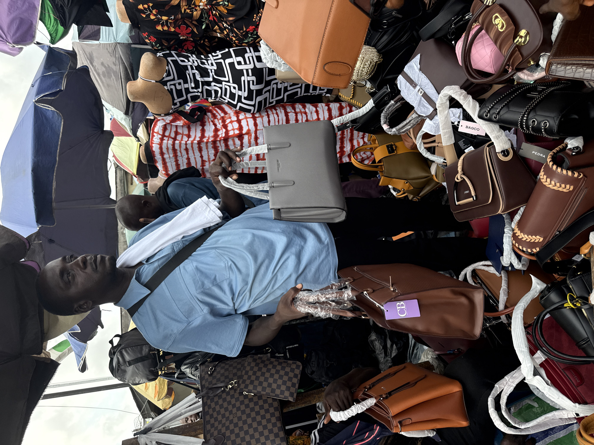 An individual in a light blue short-sleeved shirt, carrying a white towel around their neck, is positioned amid an outdoor marketplace with an array of handbags and other merchandise. The scene appears to be a retail or market setting, potentially in Lekki, Nigeria, given the style of goods and the individual's appearance. The environment includes a dense collection of bags of varying colors and materials, arranged on a platform or stall, and is partially covered by umbrellas and other overhead structures. The individual appears to be looking upwards, perhaps interacting with a customer or vendor.